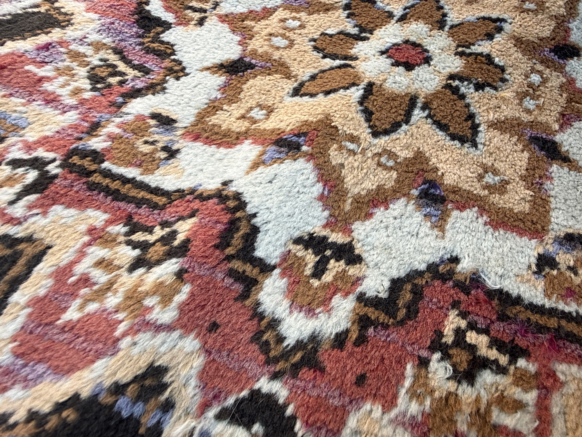 The image is a close-up, detailed photograph of a richly patterned carpet with a texture that appears soft and possibly woven. The carpet displays a complex geometric and floral design in a variety of colors, including shades of red, pink, beige, brown, black, white, and light blue. The pattern is intricate, with stylized floral elements and angular motifs. The fibers of the carpet are visible, giving it a fuzzy, tactile quality. The lighting is even, highlighting the texture and colors without harsh shadows.
The location of Okhtyrka, Ukraine, is not discernible from the provided image. The image solely focuses on the texture and pattern of a carpet, offering no environmental context or information related to a specific geographic location within a city or any other setting.