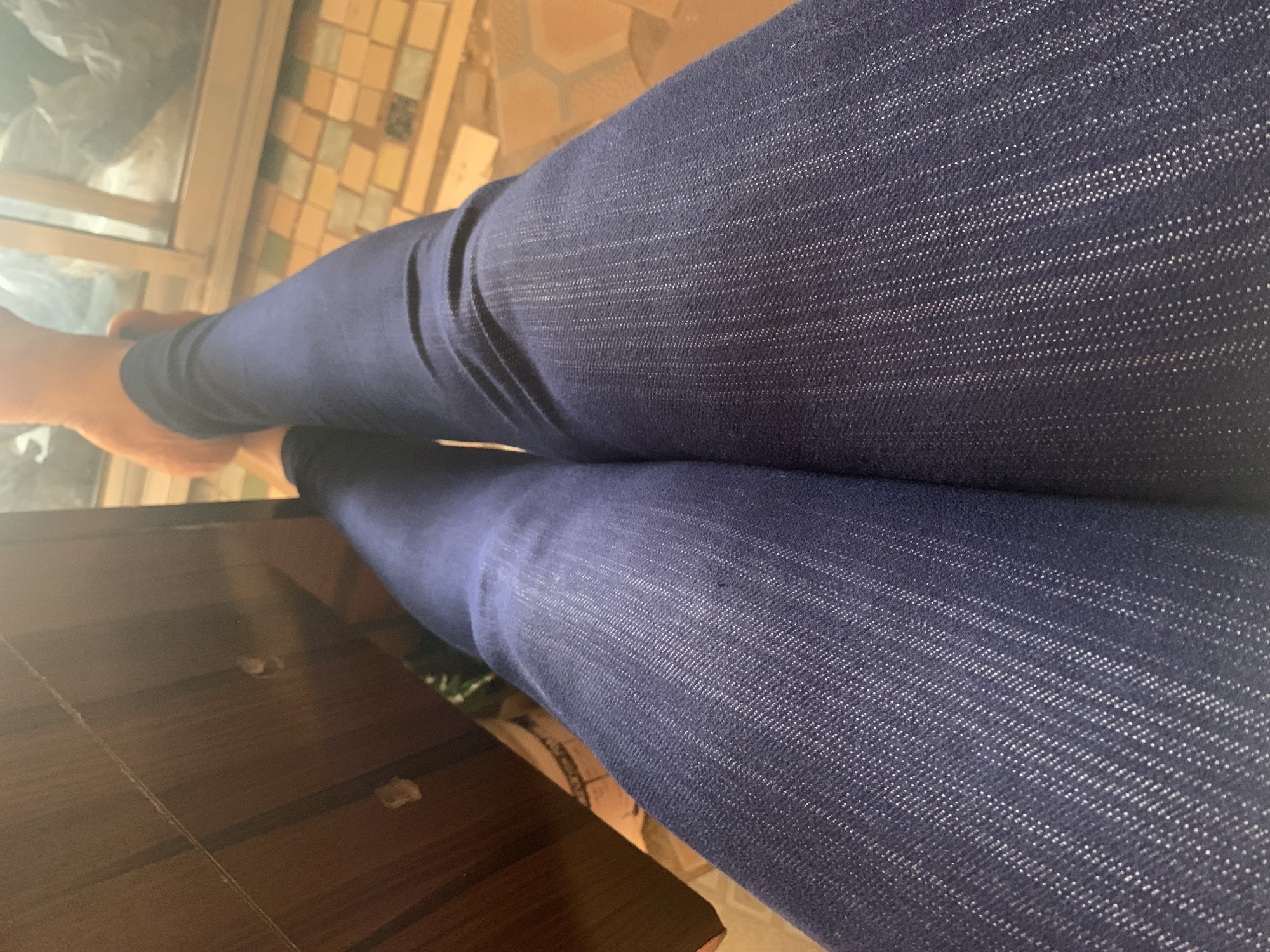 A person wearing dark blue jeans is sitting indoors. The person's bare feet are visible in the top left corner. The jeans are textured with a subtle white pattern. A wooden surface, possibly a table, is in the lower left corner. A window and tiled floor can be seen in the background. The image does not contain information to confirm the location in Sabon Tasha, Nigeria.