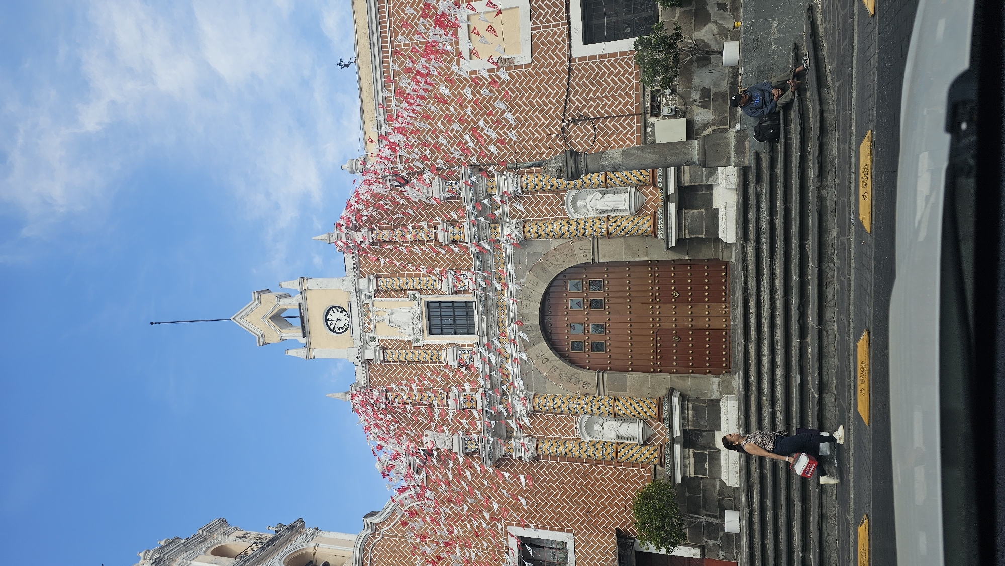 The image depicts the facade of the church of San Francisco Acatepec in Puebla, Mexico, adorned with red and white flags. The church features a brick facade, stone accents, a clock tower, and a large wooden door. On the steps leading up to the church, two individuals are present: one sitting on the steps near the bottom and another sitting near the top. The scene includes a partial view of a vehicle's window on the right side of the image.