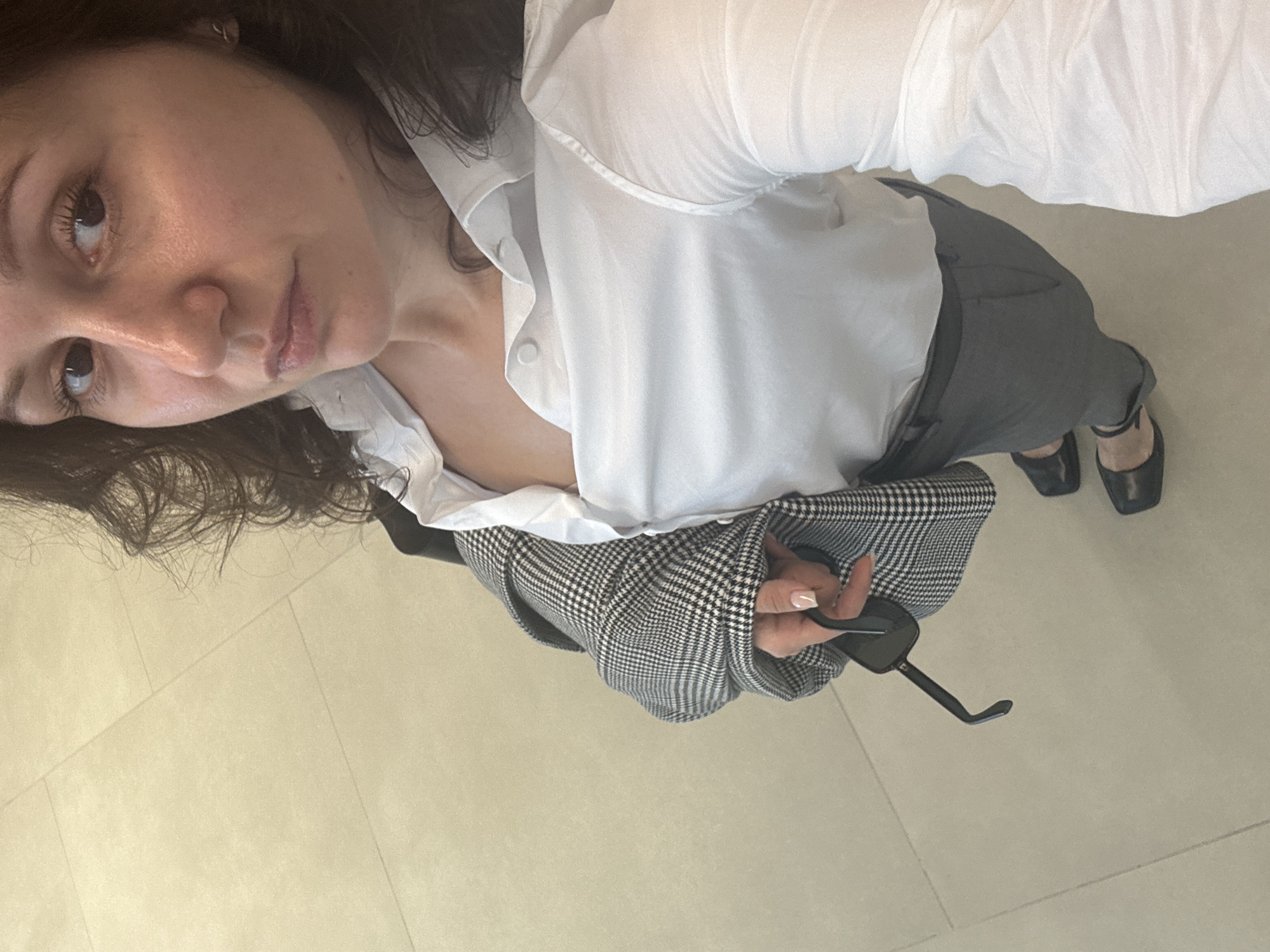 A woman with dark hair is seen from above, wearing a white button-down shirt, gray pants, and black flat shoes. She holds a pair of sunglasses in her right hand. She wears a gray and white houndstooth blazer. The location is a tiled floor. There is no specific information to identify the scene as Dubai, United Arab Emirates.