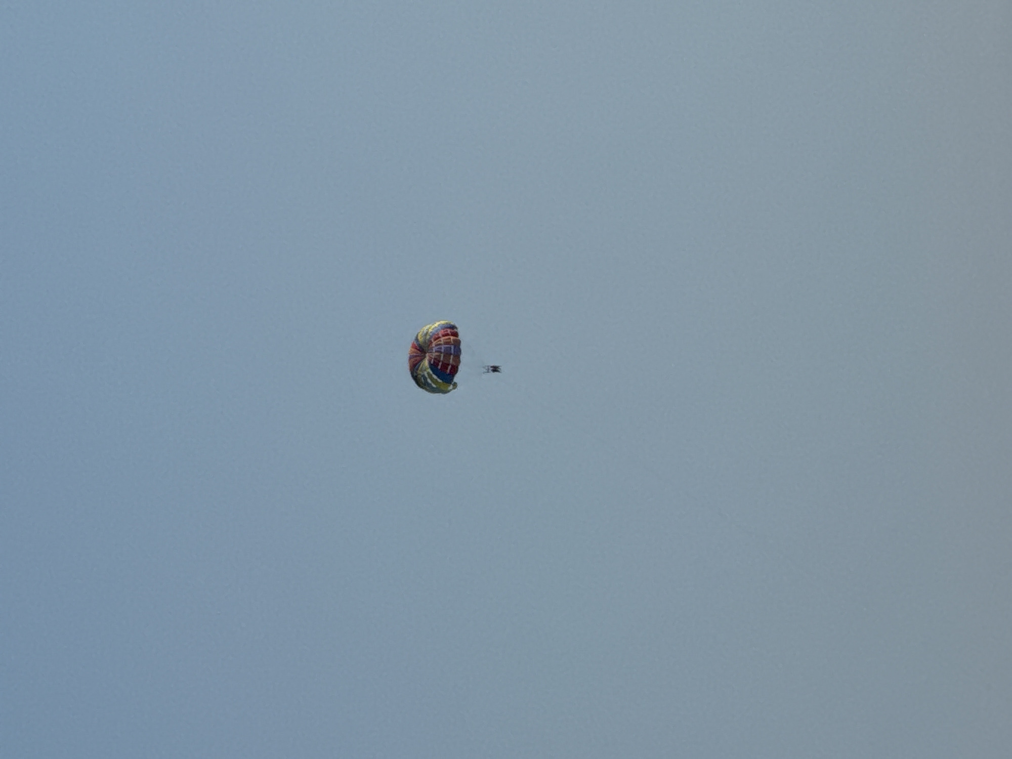 The image shows a parasailer in mid-air against a clear blue sky. The parasail is colorful, with segments of red, yellow, blue, and green. A person is suspended below the parasail, appearing as a small dark silhouette. The parasailer is positioned in the lower-left quadrant of the frame, with a vast expanse of sky above and to the right. There are no other discernible objects or features in the sky, suggesting an open-air activity. The lighting indicates it is daytime, with no clouds visible, implying fair weather. The scene is peaceful, with the focus solely on the solitary parasailer enjoying the flight. The location context of Kamenovo, Montenegro, suggests this activity is likely taking place over the sea or coastline, a common setting for parasailing. No text is visible in the image.