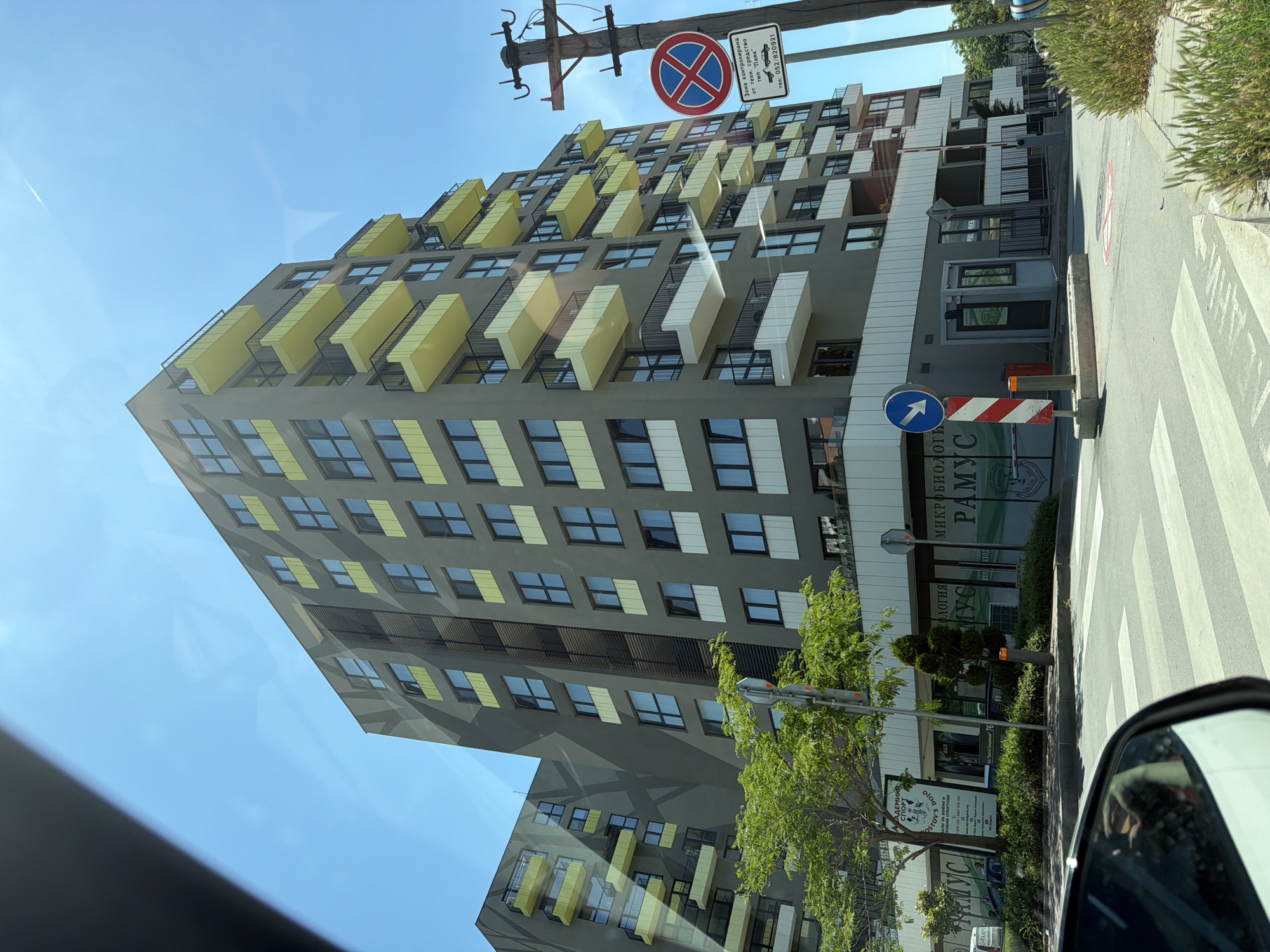 The image shows a modern, multi-story residential building with grey facade and yellow balconies, located in the city of Varna, Bulgaria. The building is situated on a street with a pedestrian crossing in the foreground. Signs for a "Dojo" and "Ramicus" are visible on the ground floor of the building, indicating commercial establishments. A traffic sign with a red circle and a blue cross, indicating no parking, is posted on a utility pole. The sky is clear blue. The scene appears to be captured from a vehicle, with a side mirror visible in the bottom left corner of the image.