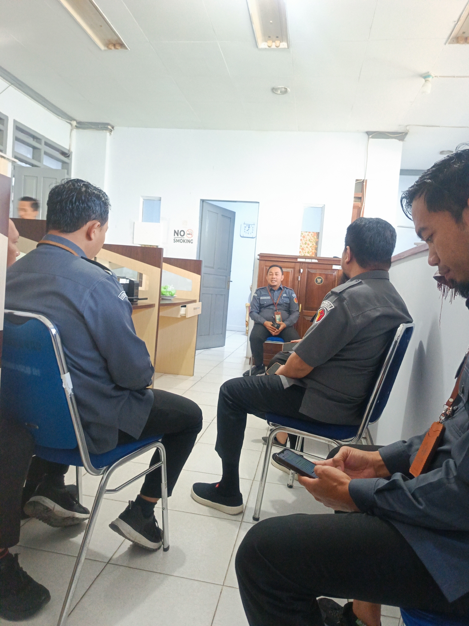 An indoor scene depicts four men in a room that appears to be an office or waiting area. Three men are seated on blue chairs, facing away from the camera. One man in the background, also seated, is facing the other three. He is wearing a dark blue uniform with a badge on his chest and appears to be speaking or presenting. The other three men are dressed in dark grey uniforms and black pants. One of them in the foreground is looking down at his mobile phone, which he is holding in his hands. Another man to his left is also looking down, his face partially visible. The room has white walls, a tiled floor, and office cubicle partitions made of light wood. A "NO SMOKING" sign is visible on the wall behind the seated men. The overall impression is a formal meeting or briefing. The specific location within the city of Gorontalo, Indonesia, cannot be determined from the visual information alone.