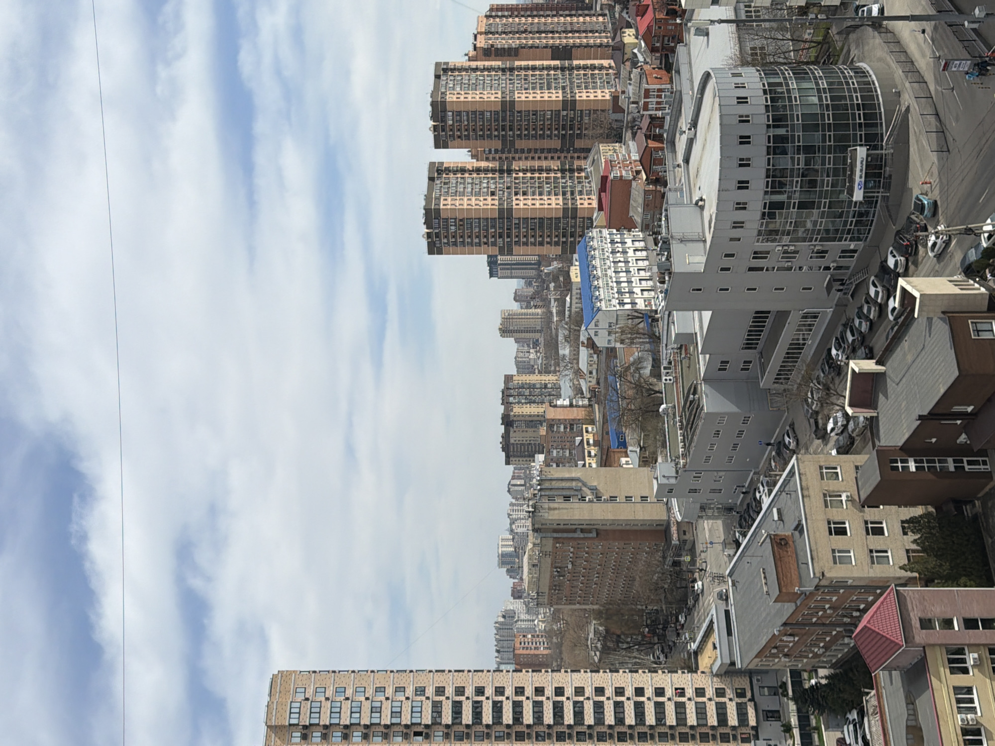 The image shows an urban landscape featuring a cluster of buildings of varying heights and architectural styles. At the top center and extending towards the left, are multiple high-rise apartment buildings, primarily in beige and brown tones, with a grid-like window pattern. To the right is a modern, semi-circular glass and concrete building, possibly a car dealership, with vehicles visible in its parking area. The middle ground consists of a mix of older, smaller buildings, interspersed with trees and roadways. The foreground features the top of a large building with regularly spaced windows. The sky is partially cloudy, with white streaks against a blue backdrop. There is a thin wire spanning the width of the frame, running across the sky. The overall impression is of a dense urban area with a mix of residential and commercial properties.
Based on the presence of a Subaru dealership, this image is likely in the city of Azov, Russia.