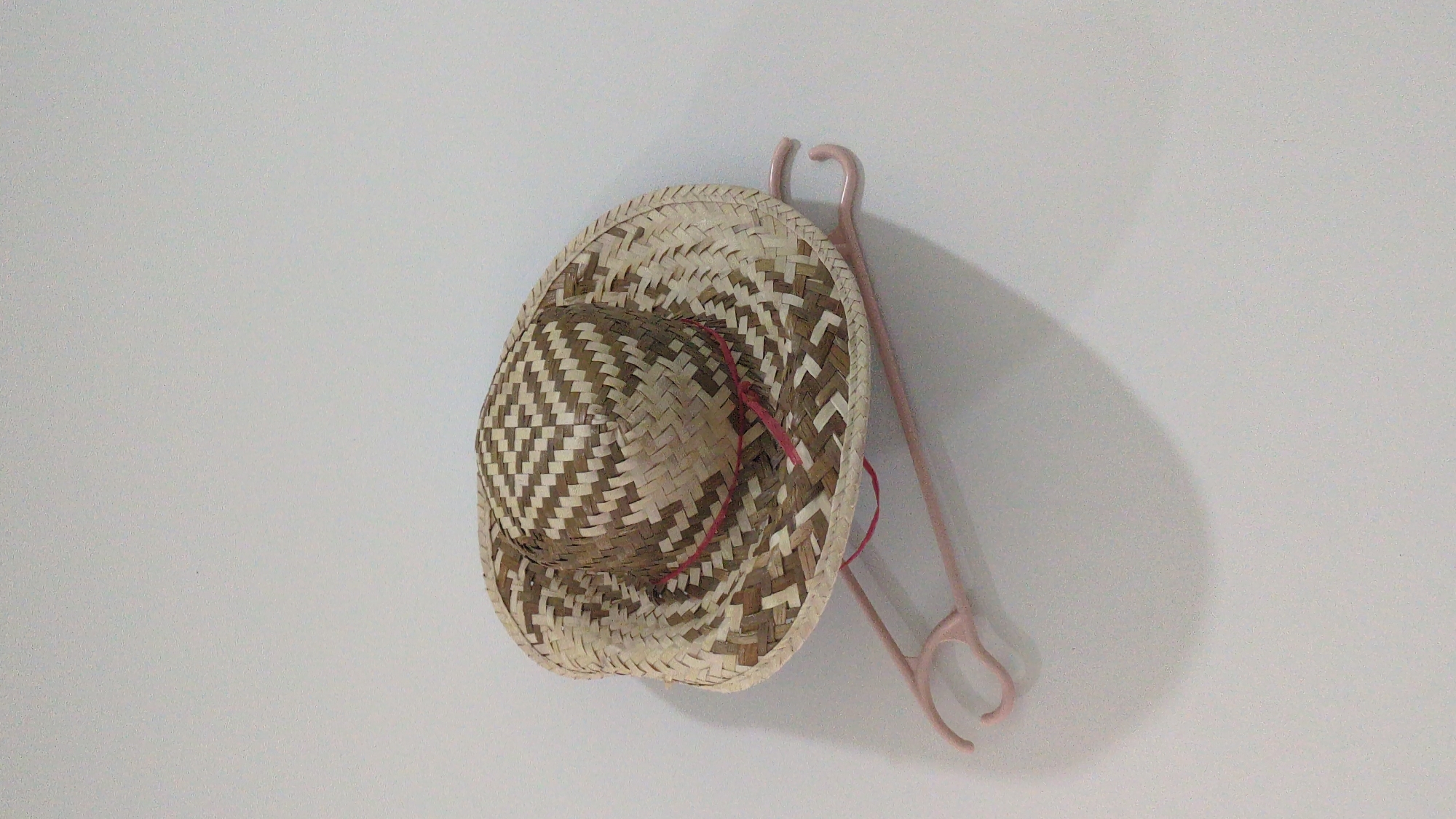 A woven straw hat with a red ribbon is hanging on a light pink hanger against a white wall. The hat is tilted, casting a shadow on the wall. The hanger is a simple, open design with two hooks. The scene is devoid of human presence and appears to be indoors. The location in Da Lat, Vietnam, is not determinable from the visual information provided.