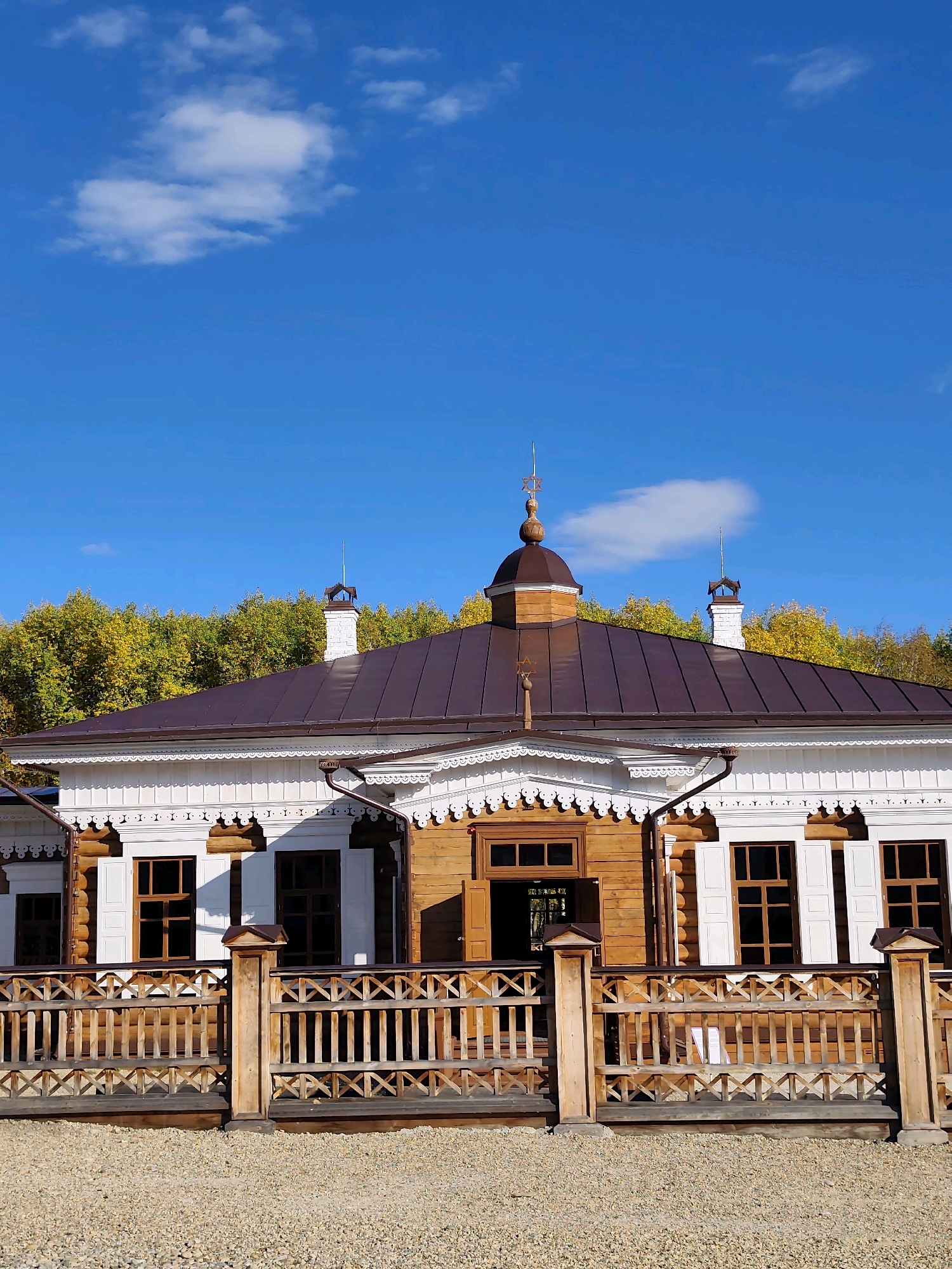 The image shows a wooden house with a brown metal roof in the village of Тальцы (Irkutsk Oblast), Russia. The house has white trim around the windows and a decorative wooden fence in front. Behind the house, there is a line of trees with yellow and green foliage, suggesting it is autumn. The sky is blue with a few white clouds. The ground in front of the fence is covered with small gravel. There are no people visible in the image.