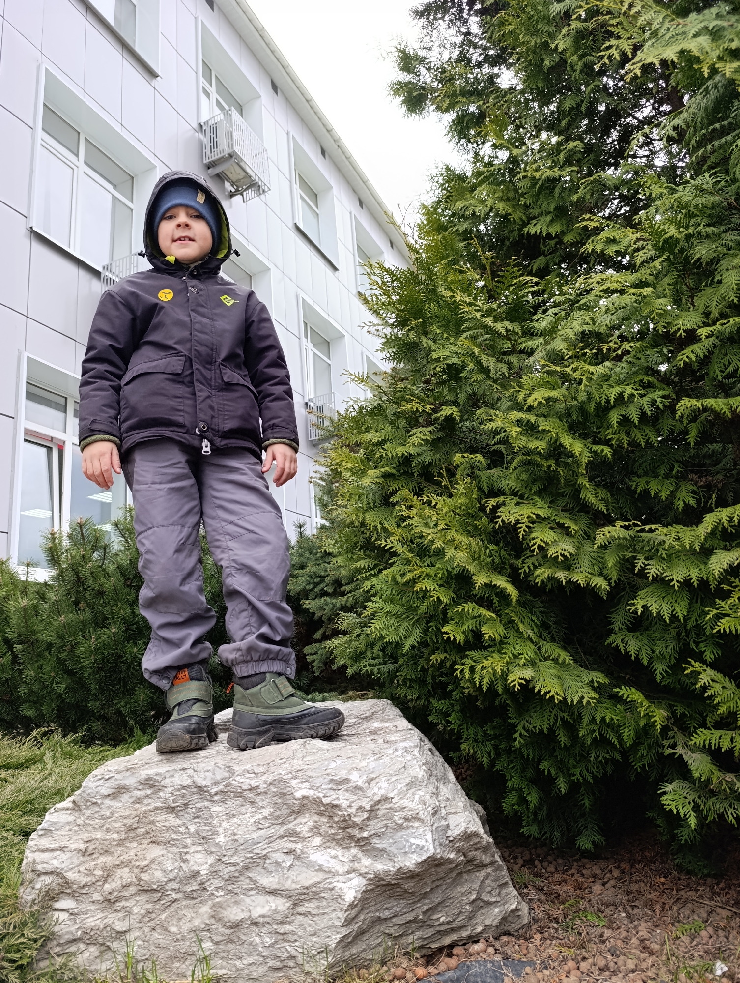 A boy stands atop a large rock in Chaikovsky, Russia. He wears a dark jacket with a hood, gray pants, and olive-green boots. Behind him is a building with gray siding and white-framed windows, as well as a large evergreen tree. The photograph is taken at eye level.