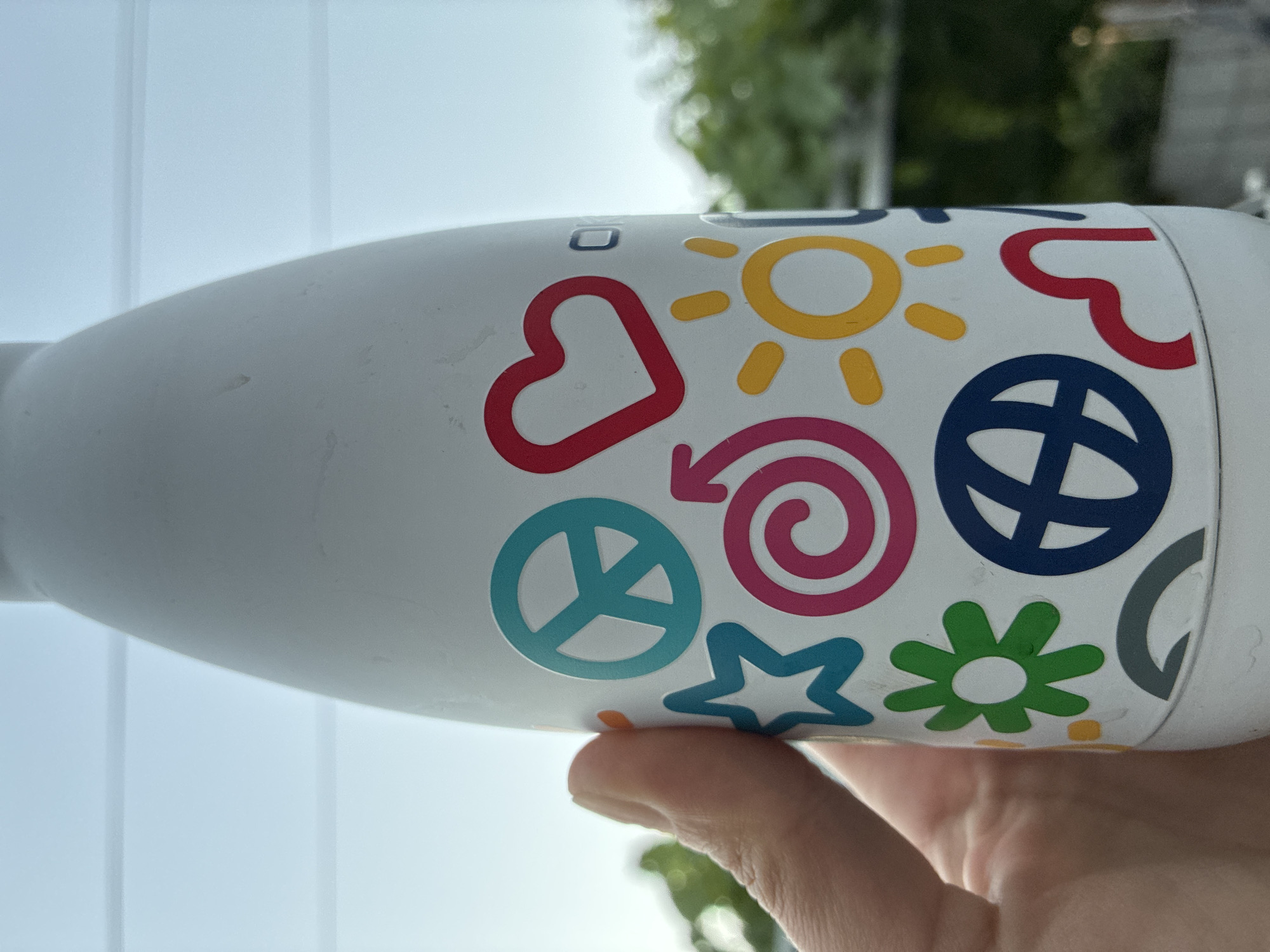 A close-up shot shows a white water bottle adorned with colorful stickers. The stickers depict various symbols such as a peace sign, a heart, a star, a spiral, a sun, a flower, a globe, and a red wavy line. A person's thumb and forefinger are visible on the left side of the bottle, holding it. The background is blurred, showing a bright sky with horizontal lines and green foliage. There is no discernible indication of the location being the city of Surami, Georgia.