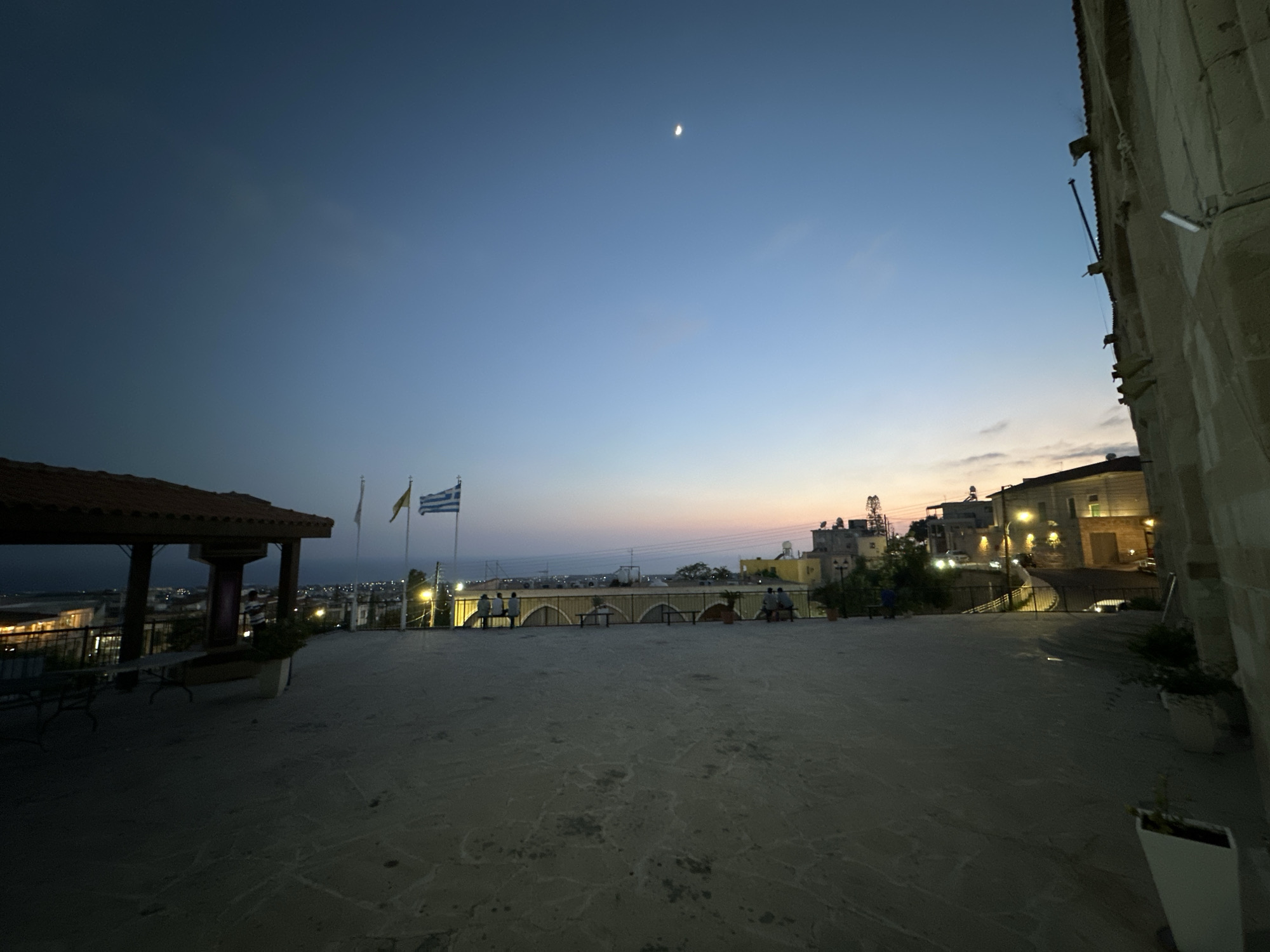 The image captures a twilight scene in Peyia, Cyprus, likely from a vantage point overlooking the town. A pale crescent moon is visible in the darkening blue sky. In the foreground, a paved area leads towards a covered structure with a brown tiled roof on the left, where tables and benches are present. Three flags, one of which appears to be the Greek flag, stand on poles behind a railing. A line of arched structures with people sitting on benches runs across the middle ground. Beyond this, the town of Peyia is illuminated by streetlights, with buildings and structures visible against the horizon. On the right side of the image, a stone building with a textured facade dominates the frame, with outdoor lighting fixtures attached to it. The overall impression is a serene evening view of the coastal town.
