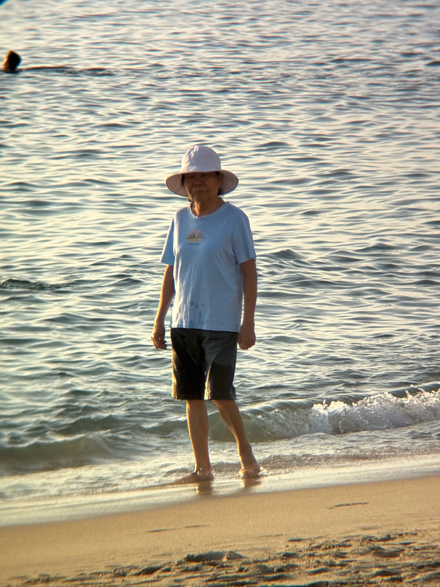 A full-shot, eye-level medium shot shows an elderly Asian woman standing on a sandy beach in Nha Trang, Vietnam. The woman is wearing a light blue t-shirt, dark shorts, and a white sun hat that covers her face. She is facing the camera, with her feet submerged in the gentle waves of the ocean. The water is clear and calm, with small ripples indicating a light breeze. The sand is light brown and appears wet near the waterline, suggesting recent wave activity. In the background, the ocean stretches out to the horizon, reflecting the golden light of what appears to be late afternoon or early morning. The sky is not visible in the frame, but the lighting suggests an outdoor setting. To the left of the woman, partially visible in the distance and out of focus, another person is swimming in the ocean. The overall impression is one of tranquility and a peaceful moment by the sea.