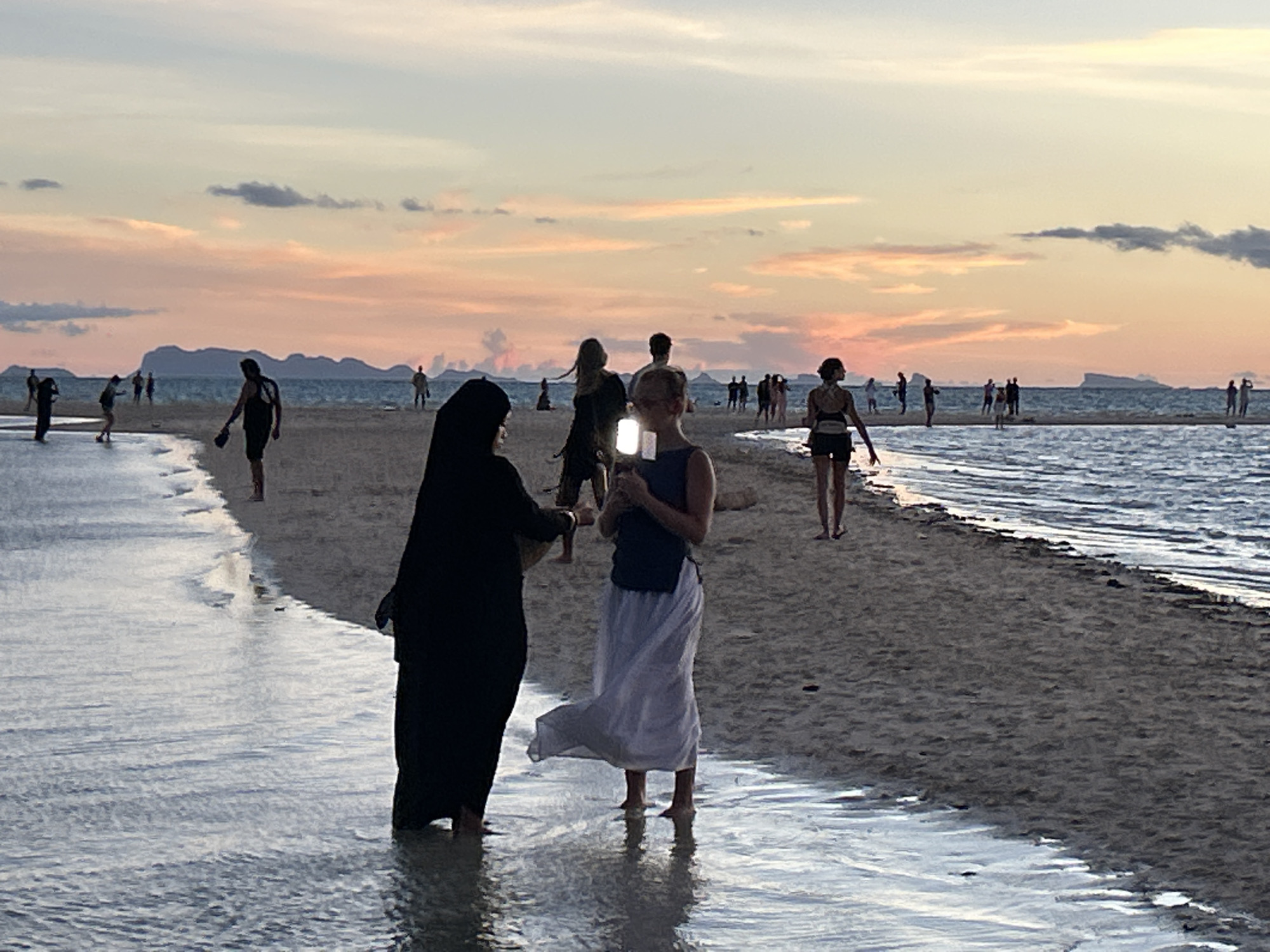 At sunset on a beach in Koh Phangan, Thailand, several people are visible wading in shallow water or walking on the wet sand. In the foreground, two women are standing. The woman on the left is wearing a black abaya. The woman on the right, facing away from the viewer, is wearing a dark top and a long white skirt, and is holding a phone up to take a photo. Behind them, more figures are scattered across the beach and in the water, silhouetted against the colorful sky. The ocean stretches to the horizon, where islands are visible as dark shapes against the gradient of pink, orange, and pale blue hues of the sunset.