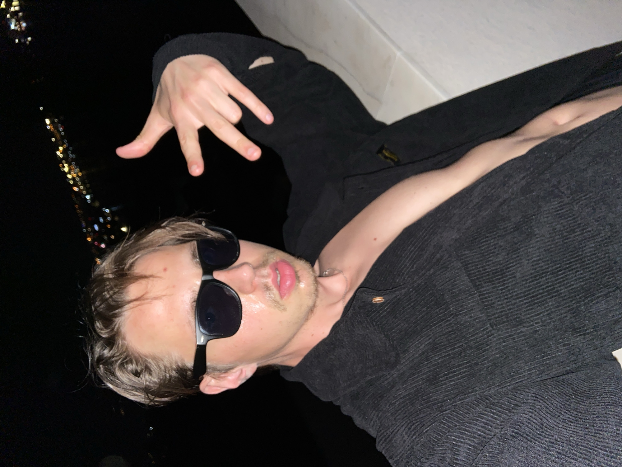 A diagonally oriented nighttime portrait features a young man with light brown hair, stubble, and prominent pink lips, wearing black sunglasses and a dark, ribbed long-sleeved shirt or jacket. He is positioned in the foreground, looking towards the viewer with a relaxed expression, partially obscured by his sunglasses. His right hand is raised, performing a gesture where the thumb and pinky fingers are extended, and the middle two fingers are bent, often recognized as a "shaka" or "hang loose" sign. A faint silver-colored chain or necklace is visible around his neck.
The background is dark and out of focus, dominated by numerous blurred lights, creating a bokeh effect. These sparkling lights suggest an urban or coastal landscape at night, seen from a distance or height, consistent with the location context of Šušanj, Montenegro. The man appears to be leaning against a light-colored, possibly white, architectural element like a wall or railing that diagonally traverses the upper right of the frame.
The scene depicts a casual, nighttime setting, likely outdoors. The time of day is clearly night due to the darkness and artificial lights. The man's pose and hand gesture convey a confident and relaxed mood. No specific weather conditions are discernible, and no text is visible in the image.