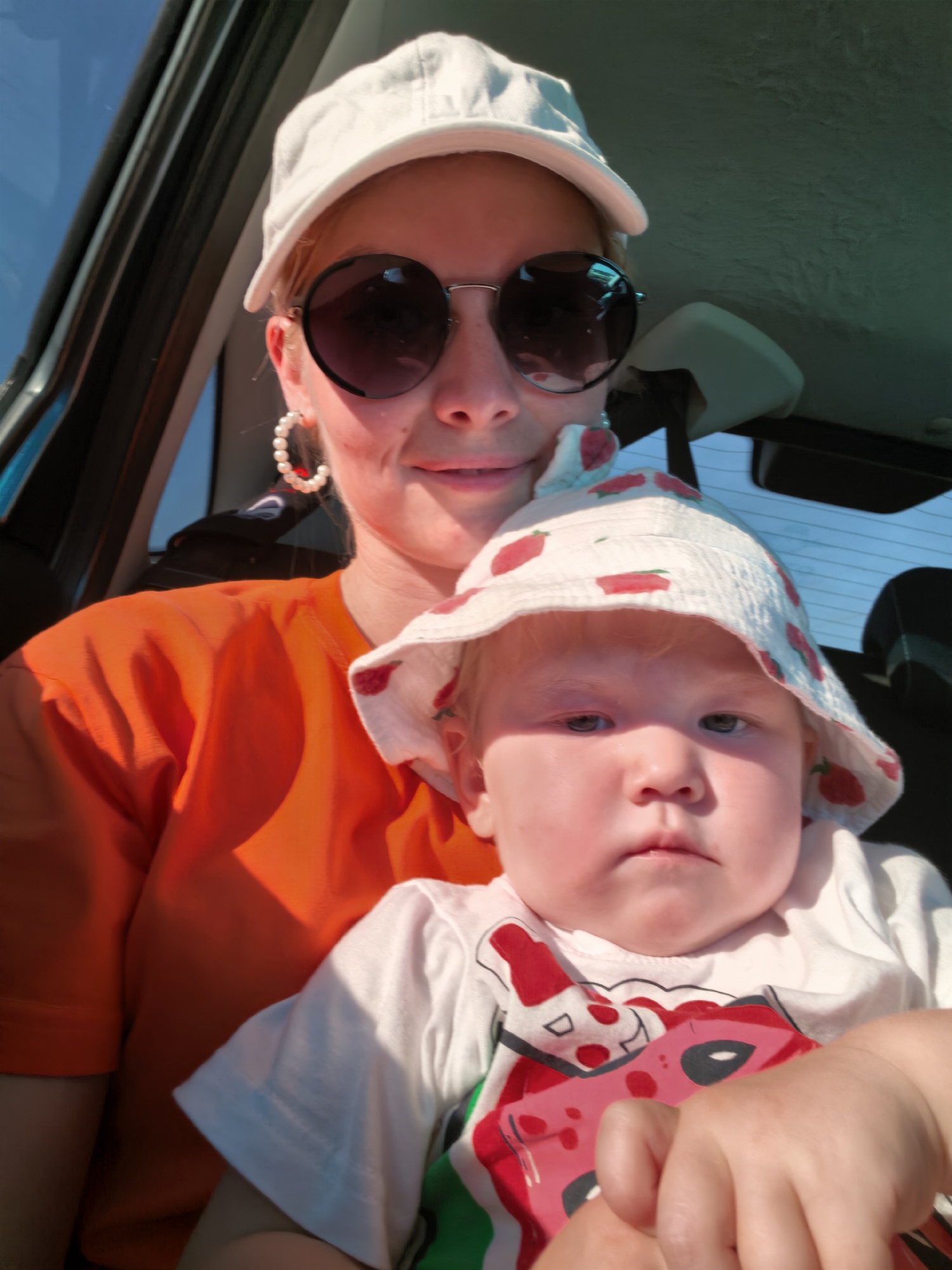 A woman and a baby are in the back seat of a car, likely in Karaiganovo, Russia. The woman is wearing a white baseball cap, large round sunglasses, and an orange t-shirt. She has on large pearl hoop earrings. The baby is wearing a white bucket hat with red strawberry prints and a white t-shirt with a cartoon character on it. The baby is being held by the woman. The image is taken from a selfie perspective, with the sun creating harsh shadows and highlights. The background shows the interior of a car, including the back window with sunlight streaming through it.