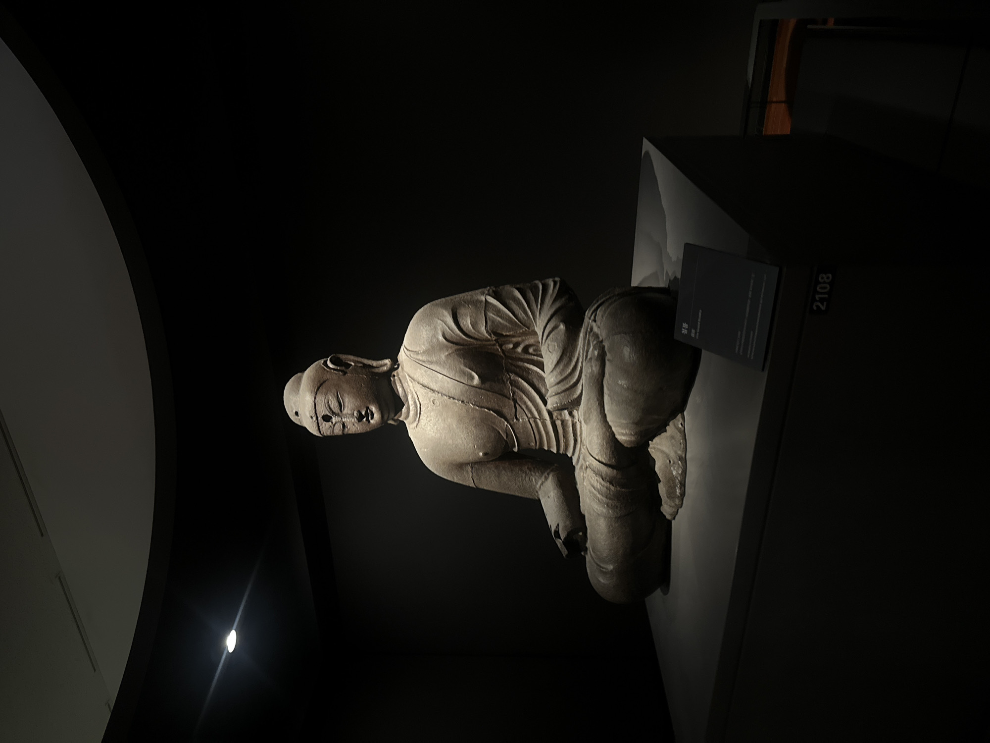 A seated stone statue of Buddha is displayed in a dimly lit museum. The statue is positioned on a dark, rectangular pedestal with a small information plaque to its right. The background is dark, with the only illumination coming from a spotlight above and to the left of the statue, casting dramatic shadows. The statue depicts Buddha in a meditative pose, with legs crossed and hands resting on his lap. The carving is detailed, showing the folds of the robe and the serene expression on Buddha's face. The number "2108" is visible on the pedestal below the plaque. This scene is likely within the National Museum of Korea, located in Yongsan-gu, Seoul, South Korea, given the typical presentation of artifacts in such institutions.