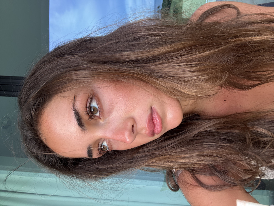 A young woman with light brown hair and olive skin gazes at the camera. Her hair is wavy and long, framing her face. Her eyes are almond-shaped, and her lips are full and glossy. She appears to be outdoors, with hints of sky and buildings in the background. The overall composition suggests a casual, natural snapshot.