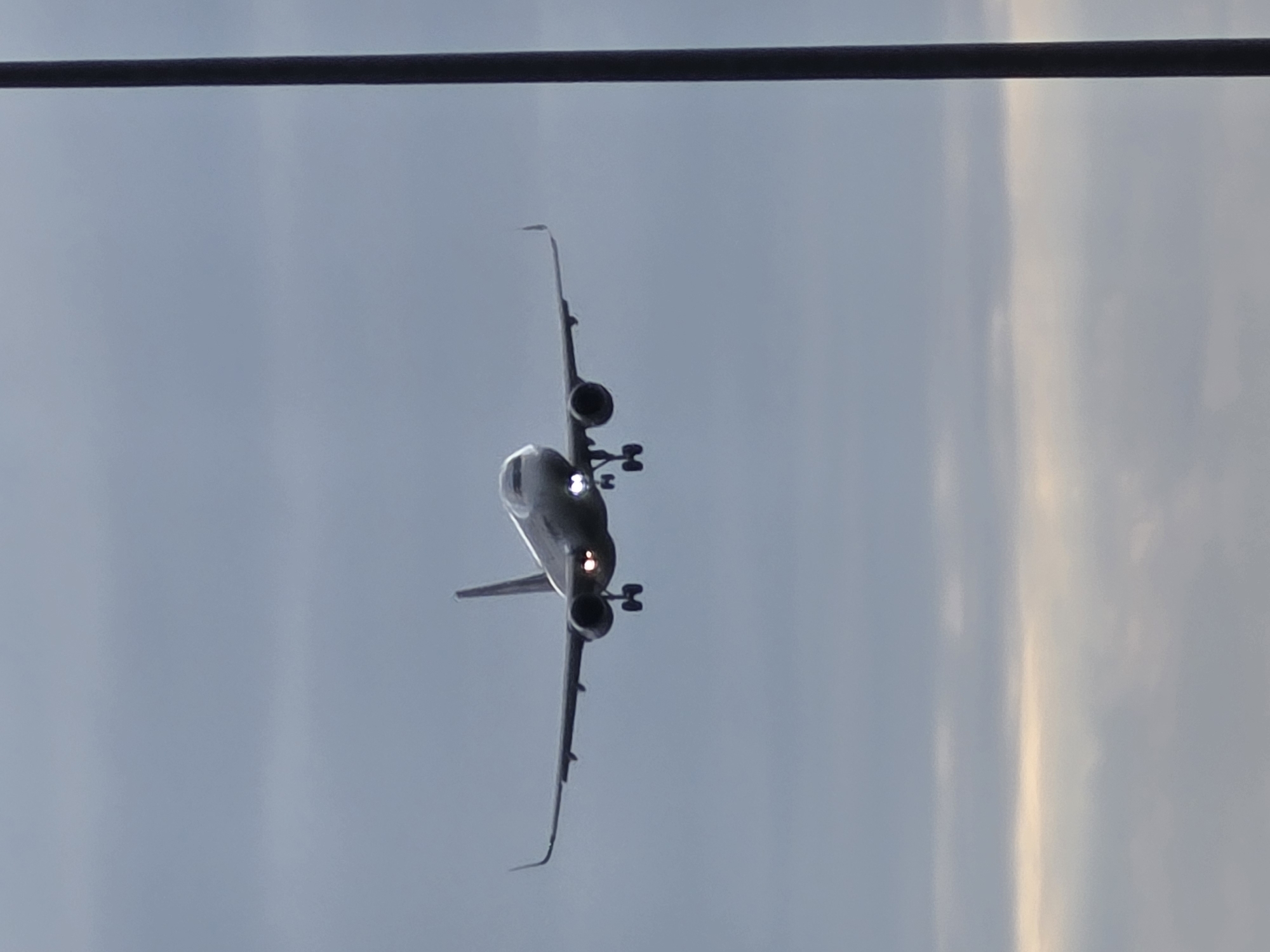 An airplane is in flight, appearing to be on a landing approach, against a cloudy sky. The aircraft is a passenger jet, with its landing gear extended. The body of the plane is silver, and the wings are angled upwards at the tips. The lights on the front of the plane are illuminated. A dark vertical pole, possibly a lamppost, is present in the right side of the image, partially obscuring the view of the sky. The sky is overcast with streaks of lighter clouds and a faint band of light near the horizon. The image appears to have been taken in Kelsterbach, Germany, likely near Frankfurt Airport, given the proximity of the airport to the city and the nature of the subject.