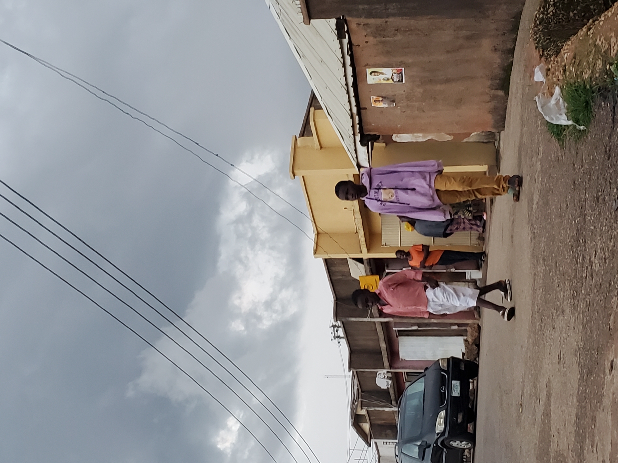 In the city of Jos, Nigeria, under a cloudy sky, two young boys walk down a paved street. The boy on the left wears a salmon-colored collared shirt and white shorts. The boy on the right is dressed in a light purple hooded sweatshirt with a teddy bear graphic and brown trousers. A black van is parked to the left of the frame, and various buildings line the street on both sides, some appearing to be shops with advertisements on their walls. The ground is asphalt, with some debris, including plastic bags, visible on the side of the road. Other people are present in the background, some walking and some standing near the buildings. Power lines are strung across the sky overhead.