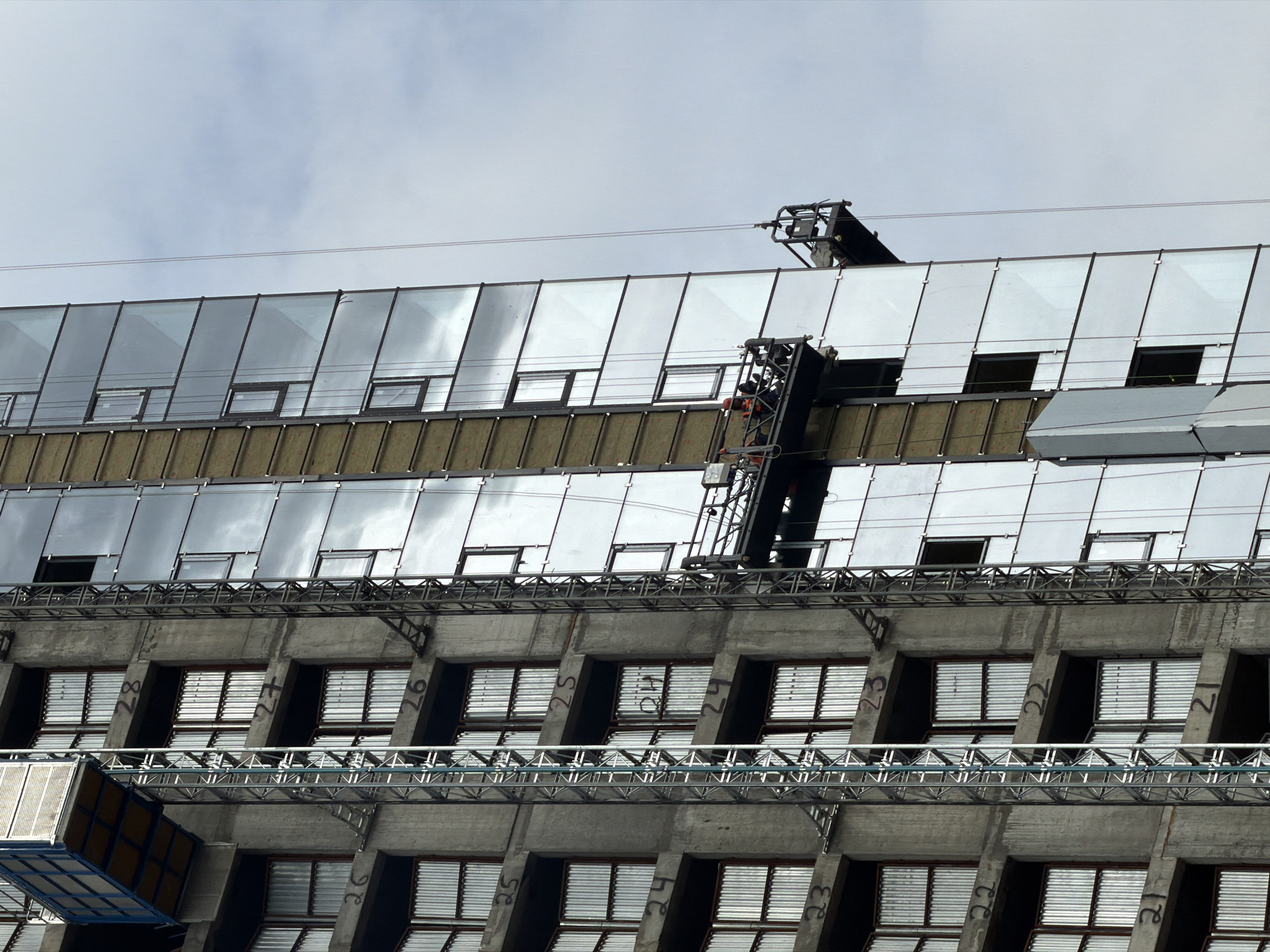 A construction worker operates a hydraulic lift attached to a large building. The building has a modern facade featuring rows of glass panels and concrete structures with many windows. The worker, wearing an orange safety vest and a hard hat, is positioned high up on the lift, appearing to be engaged in maintenance or repair work. The sky is overcast, suggesting a cloudy day, and there are no strong indications of the time of day. The scene appears to be an outdoor construction site. There is some visible text on the concrete columns, including numbers such as "28", "27", "26", "25", "24", "23", "22", and "21". The overall impression is one of ongoing construction or renovation.