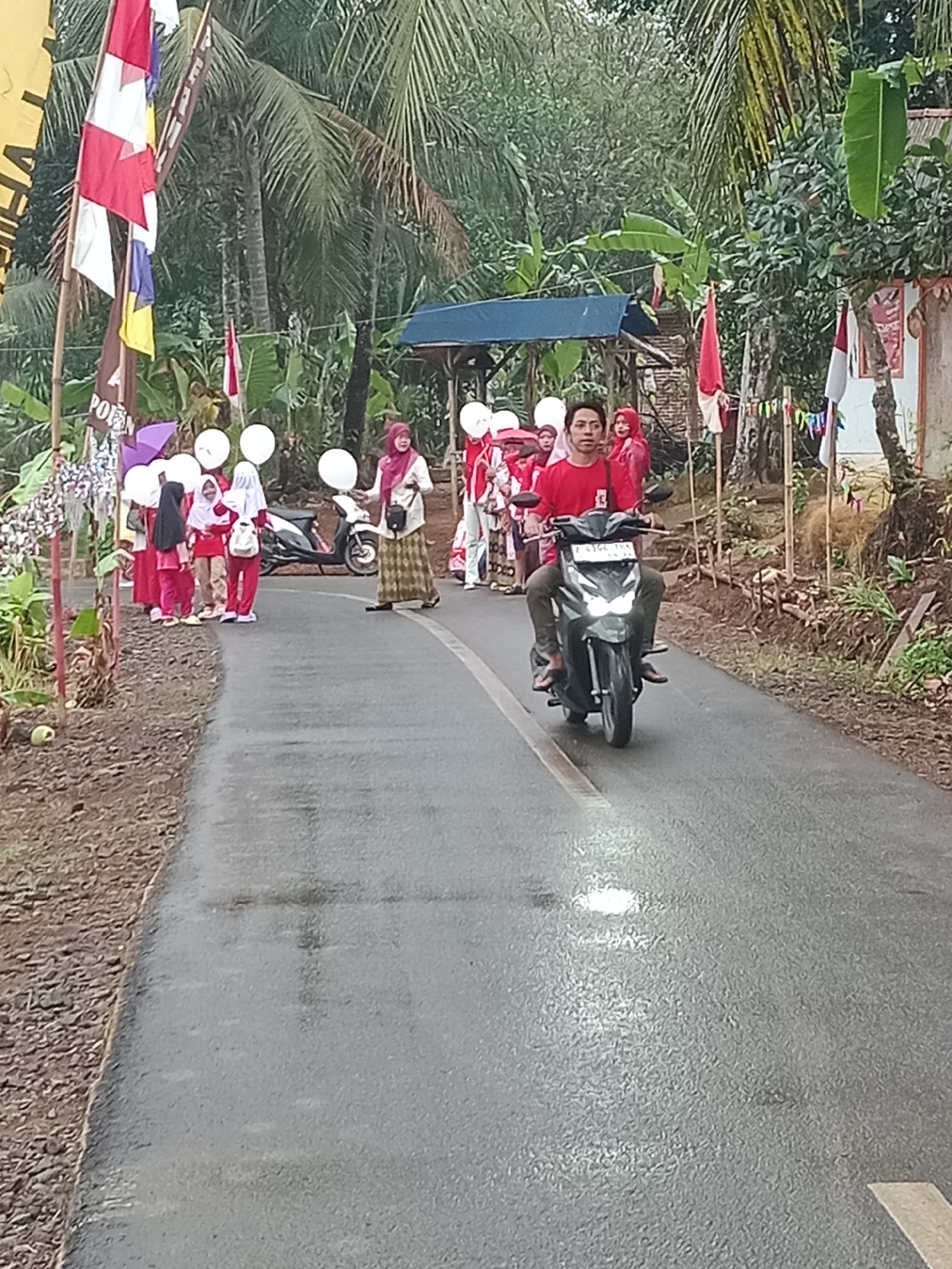 In Ciharalang, Indonesia, a group of children, some wearing red and white uniforms, participate in a procession along a paved road. They are holding white balloons. A young man rides a motorcycle in front of them, wearing a red shirt. The procession is taking place amidst lush green vegetation, including palm trees and banana trees. Red and white flags are visible, suggesting a festive or patriotic event. The road appears wet, possibly from recent rain. The scene is set against a backdrop of rural Indonesian landscape with some simple structures.