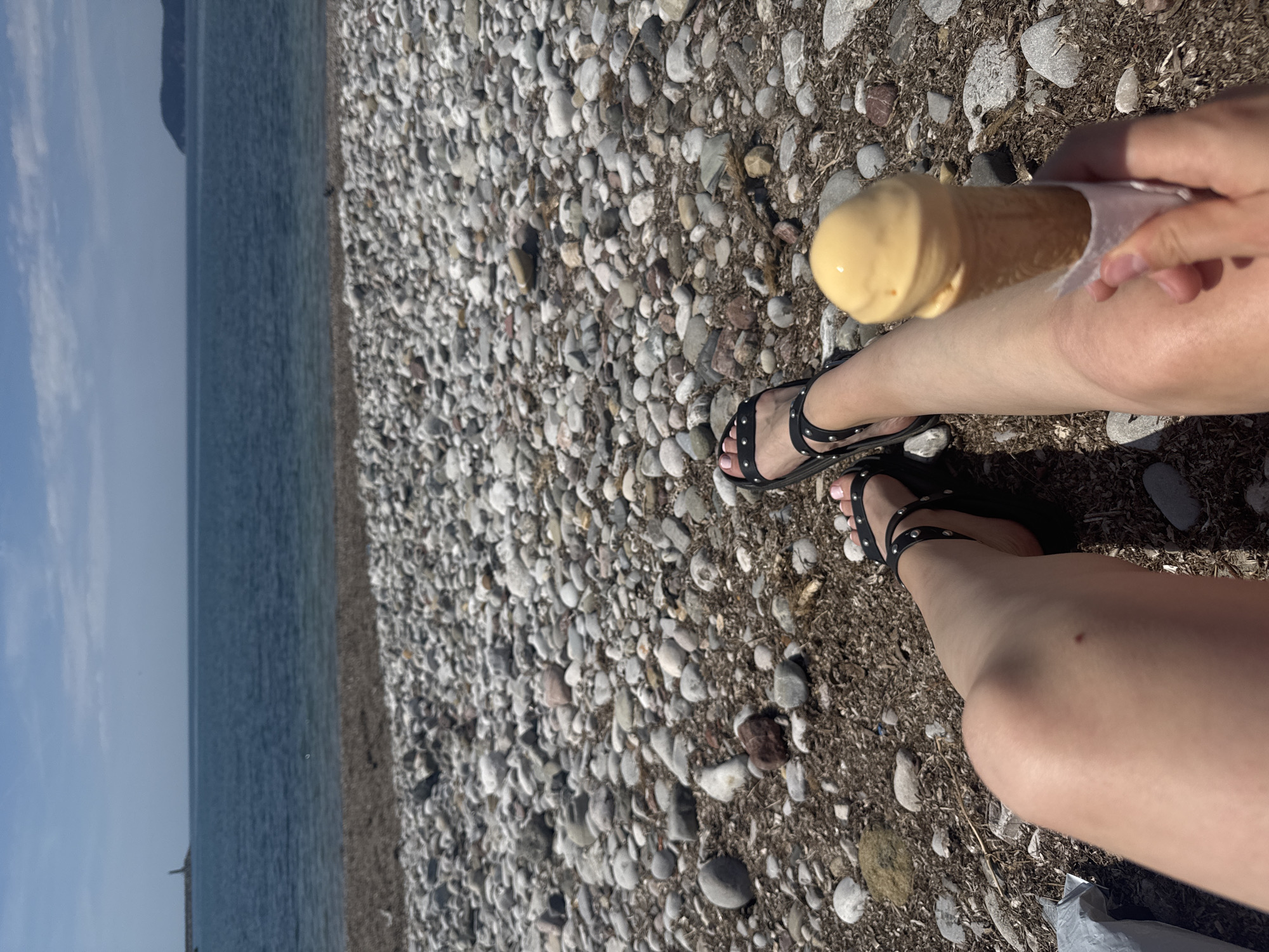 This sunlit image captures a first-person perspective of a person relaxing on a pebble beach in Bar, Montenegro. The person's legs, wearing black sandals with stud details, are extended onto the coarse, grey and brown pebbly ground. In the foreground, a hand holds an ice cream cone topped with a scoop of light yellow-orange ice cream, which appears slightly melted. To the left, the calm, blue-grey sea meets the horizon under a bright, clear sky. A faint, dark landmass or mountain is visible in the distance across the water. The scene evokes a leisurely summer day by the coast. No text is visible in the image.