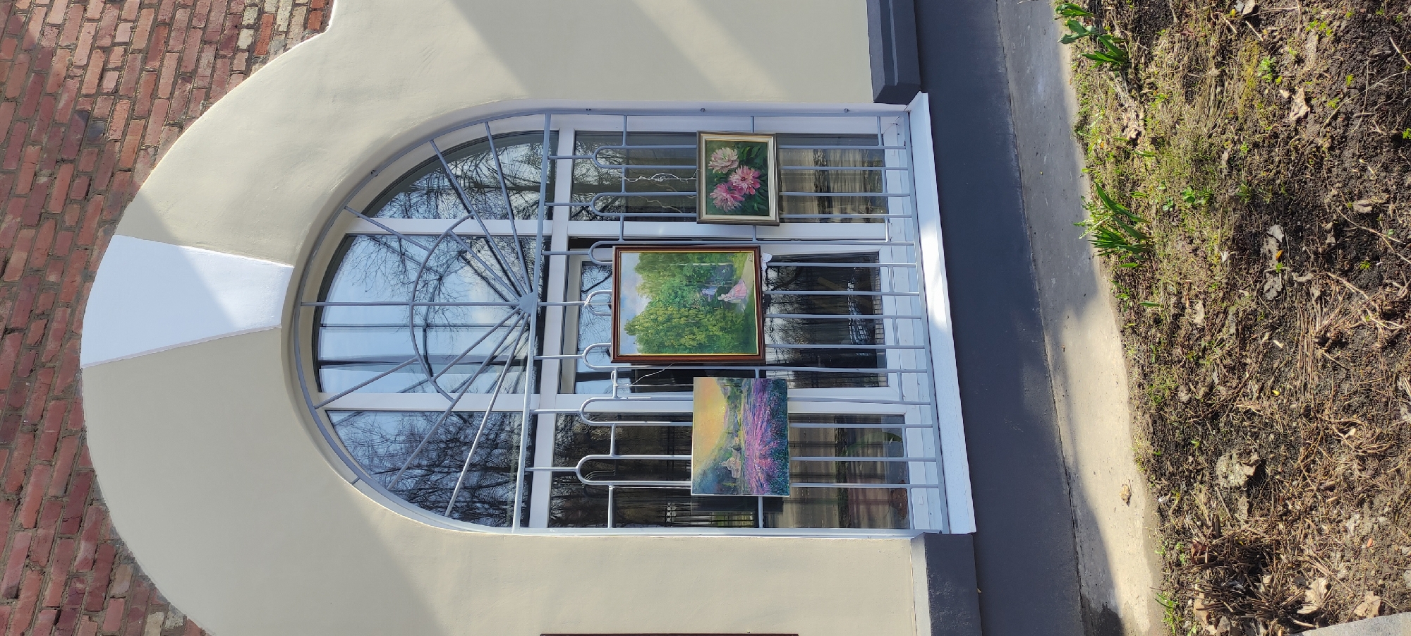 The image shows a partially visible rounded window of a building in Fryazino, Russia. The window is covered with a decorative metal grid. Three paintings are hung on the grid: one with flowers at the top, another with a landscape in the middle, and a third with a lavender field at the bottom. The window frame is off-white, with a white geometric section on the left side. The building is made of brick on the left side of the window and off-white stucco around the window itself. To the right of the window is a concrete ledge, and next to that is an area with dirt, grass, and some small plants. Bare tree branches are visible through the window.