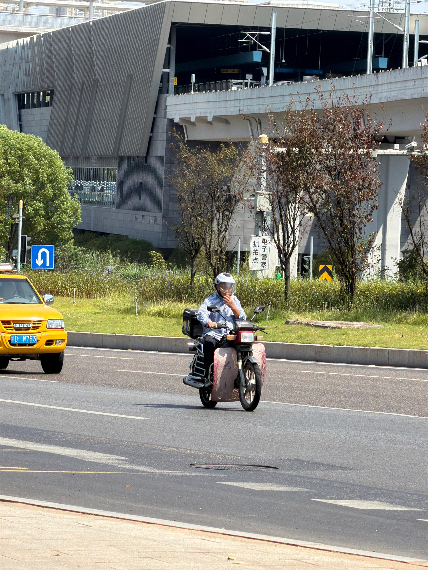 A person wearing a checkered shirt and a white helmet rides a scooter on a paved road in Jiangjin, China. The scooter has a pink, patterned shield on the front. The person is looking down and to the right, possibly at their phone, which is held near their face. To the left of the scooter, a yellow taxi with the license plate "D-LW753" is partially visible. In the background, there is a large, modern building with a corrugated gray facade, a tree-lined median with purple flowers, and traffic signs, including a blue U-turn sign and a white sign with Chinese characters that appears to indicate a traffic camera or police presence. The overall scene depicts a typical urban street environment with traffic and infrastructure.