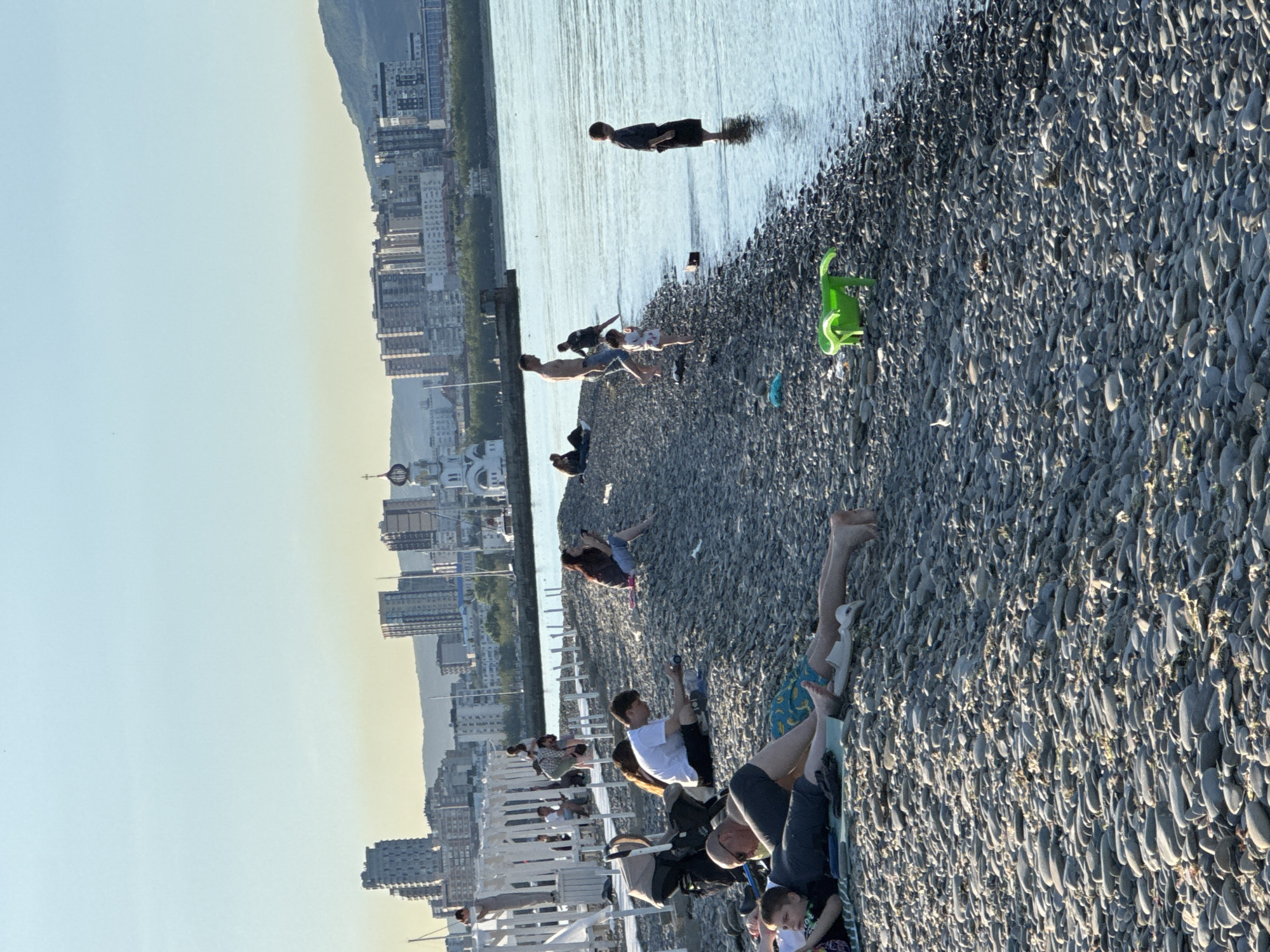 This image captures a vibrant late afternoon or early evening scene on a pebble beach in Novorossiysk, Russia. The sky, clear and bright, transitions from a light blue overhead to a warm, soft yellow hue near the distant horizon, indicative of the golden hour.
The foreground and mid-ground are dominated by a dark gray pebble beach, teeming with people enjoying the leisure time. Numerous individuals, including adults and children, are scattered across the beach. Many are seen sitting or lying down on the pebbles, some on mats or towels, basking in the sun. A bright green plastic toy, resembling a dinosaur, lies on the beach. Several people are wading in the calm, reflective water, with one person standing waist-deep further out.
To the left of the frame, a white pier or boardwalk extends, with more individuals seated and standing, contributing to the lively atmosphere. A baby stroller is also visible near the pier.
The background features a sprawling cityscape with various buildings and high-rises. Distinctive white walls and golden domes of what appears to be a Russian Orthodox church or cathedral rise above the urban landscape. Behind the city, undulating hills or mountains are silhouetted against the bright sky. The overall mood is one of relaxation and enjoyment on a sunny day by the sea. No discernible text is visible in the image.