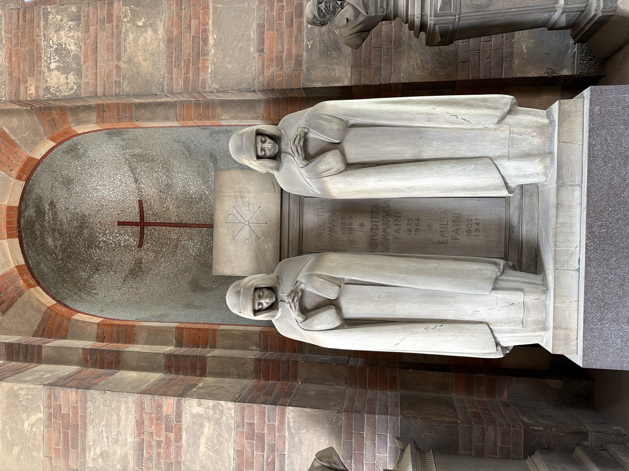 This image shows the Faini tomb in the Monumental Cemetery of Milan, Italy. The tomb features two white marble statues of women in simple robes, positioned horizontally within a recessed alcove. The alcove is lined with a mosaic of small, reflective tiles, with a red cross and another symbol etched into the mosaic behind the statues. The tomb's base is engraved with the names "CARLOTTA SOMMARVGA FAINI," "GIVDITTA SOMMARVGA FAINI," and "EMILIA FAINI," along with their birth and death years. The surrounding architecture consists of brick and stone, indicating a historical or monumental setting.