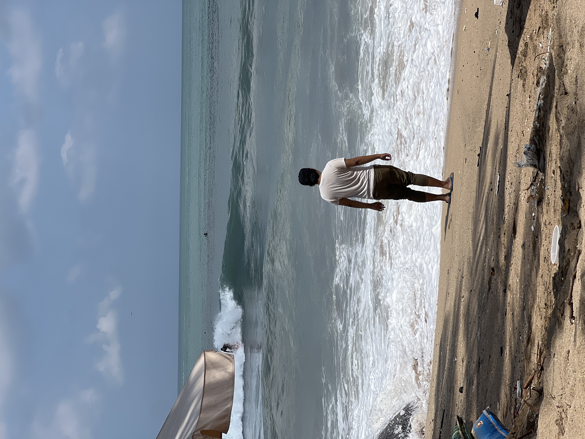 The image depicts a beach scene in Kuta, Indonesia. In the foreground, a man wearing a white t-shirt and brown pants stands facing the ocean. His back is to the camera. The beach is sandy, with some debris scattered on the ground. To the left, a portion of a large umbrella is visible. The ocean features moderate waves, and a surfer is actively riding a wave in the mid-ground. The sky is partly cloudy, with a hint of blue. The overall impression is one of a typical beach day with some activity in the water.
