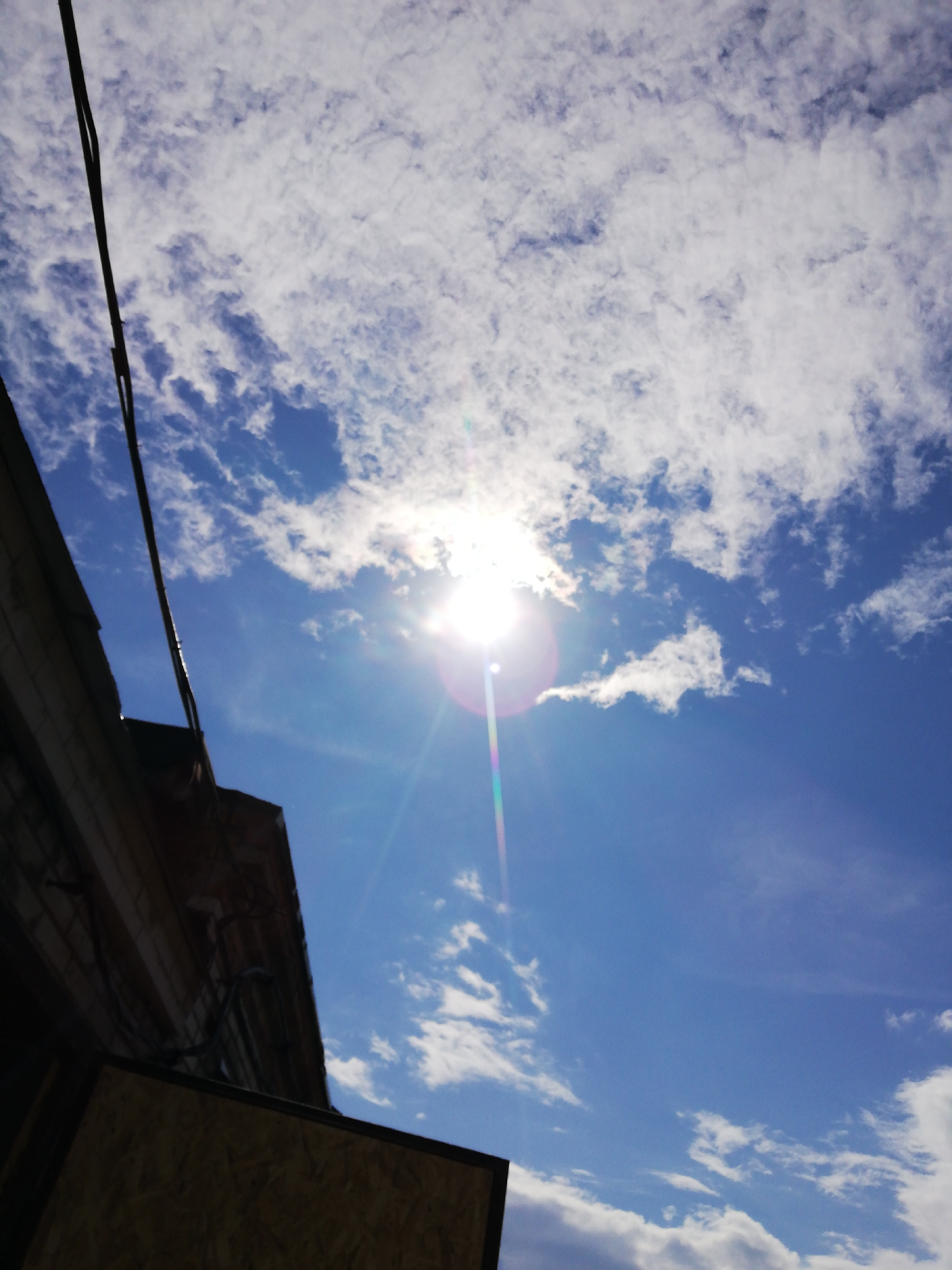 The image captures a bright, sunlit sky over a city environment, likely Tosno, Russia, as indicated. The sky is a vivid blue, dotted with scattered, wispy white clouds. The sun is prominently visible, creating a lens flare that casts a rainbow-like streak across the sky. In the lower left portion of the frame, a building's facade is visible, appearing to be constructed with light-colored bricks. A dark, thin cable runs vertically along the left edge of the image, obscuring a portion of the building and the sky. Below the building, a section of a wooden structure, possibly plywood, is visible. The overall impression is of an outdoor scene on a clear, sunny day.
