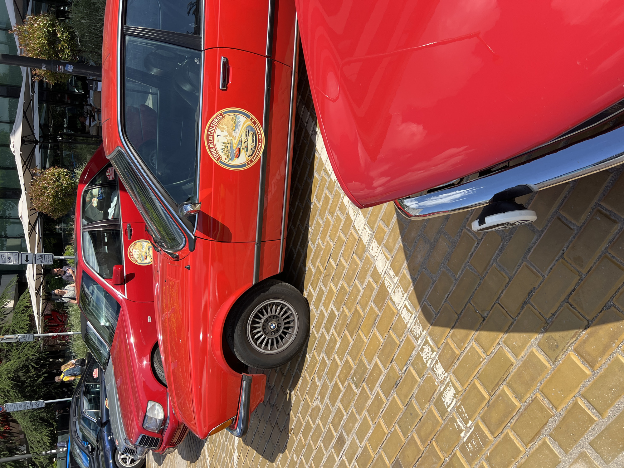 The image depicts an outdoor urban scene in Sofia, Bulgaria, identified by visible Bulgarian text ("АБОНАМЕНТ," "ЗА КЛИЕНТИ") on street signs in the background. Multiple classic red vehicles are parked on a yellowish-brown brick-paved surface, which features partially visible white parking lines.
In the immediate foreground, the shiny red hood and chrome bumper of a vehicle are partially visible at the bottom right. Directly behind this, a red two-door coupe, equipped with multi-spoke alloy wheels (bearing a BMW logo on the hubcap), is parked at a slight diagonal. This coupe features a large circular decal on its driver's side door, displaying "TOUR DE LAS CULTURAS" at the top, "BARCELONA - ISTANBUL" at the bottom, and illustrations of classic cars and a landscape in the center, numbered '1'.
Behind the red coupe, a red Mercedes-Benz sedan is parked. This vehicle also carries a similar "TOUR DE LAS CULTURAS" circular decal, numbered '10'. Further into the middle ground, additional cars are partially visible, including a dark blue convertible and a silver sedan.
The background consists of a modern building with extensive glass windows reflecting the environment. An outdoor seating area for a cafe or restaurant is set up in front of the building, featuring several white rectangular umbrellas and tables. People are present: an individual in a yellow shirt with a backpack is seen walking on the left, two men are standing near the cafe area, and another person is seated at an outdoor table. Multiple street signs on poles, along with a street light, are distributed across the background. Large green trees are visible above the building, complemented by smaller ornamental bushes in front. The scene is brightly lit by direct sunlight, casting distinct shadows on the pavement and vehicles.