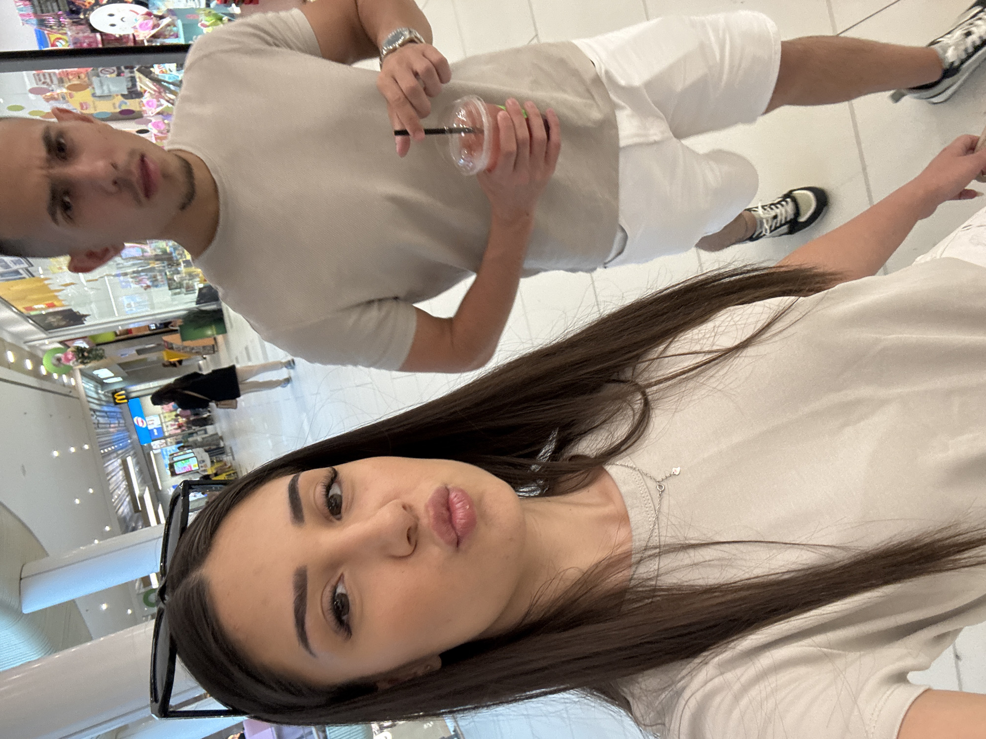 An indoor selfie captured at a shopping mall in Plovdiv, Bulgaria, depicts a woman in the foreground and a man behind her.
The woman, positioned on the left side of the frame, has long, dark, straight hair parted in the middle, extending over her shoulders. She wears a light beige or off-white short-sleeved t-shirt and a delicate silver-colored necklace with a small, possibly clover-shaped pendant. Her dark eyebrows are well-defined, and she has visible false eyelashes. Her lips are pursed in a "duck face" pose, directed towards the camera. Black sunglasses are perched on top of her head. Part of her lower body, dressed in white shorts or a skirt, is visible at the bottom. Her right hand is partially visible at the bottom right.
The man stands slightly behind and to the right of the woman. He has short, dark hair with faded sides and light facial stubble. He wears a light beige short-sleeved t-shirt and white shorts. A silver-toned watch with a round face is visible on his left wrist. In his right hand, he holds a clear plastic cup with a dark straw, containing a reddish-orange beverage. He looks towards the camera with a neutral to slightly serious expression. His footwear consists of dark-colored sneakers with white and possibly light beige accents, with one foot visible stepping forward.
The background reveals a brightly lit, spacious shopping mall walkway with a light-colored tiled floor. Large, white, slanted pillars are prominent in the upper left. Storefronts line both sides of the walkway; on the right, a distinctive yellow "M" logo, consistent with McDonald's, is visible, along with a colorful store displaying various items and a white, sad-faced balloon or decoration. Other blurred individuals are visible walking in the distance. The overall lighting is bright and even, characteristic of indoor commercial spaces.