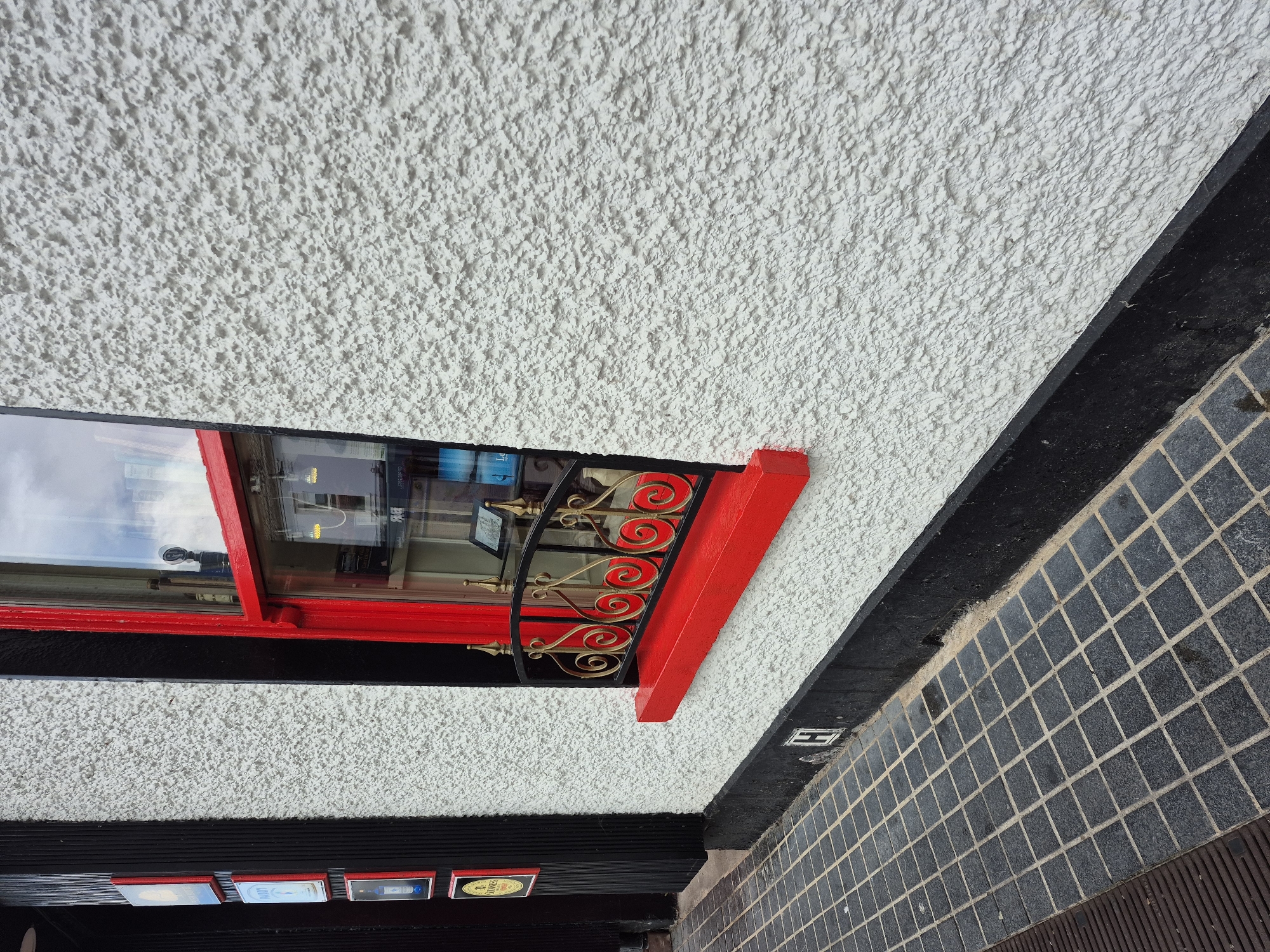The image depicts the exterior of a building in Carrick-on-Shannon, Ireland. A white, rough-textured wall with a bright red window frame is a prominent feature. The window has a decorative black metal grate with gold accents in front of it. Below the window, a red windowsill protrudes. To the left of the wall, a black vertical structure with several framed square signs, some with red borders, is visible. The ground in front of the building is paved with dark grey square tiles, and a metal grate is partially visible on the left. A black plinth runs along the base of the white wall.