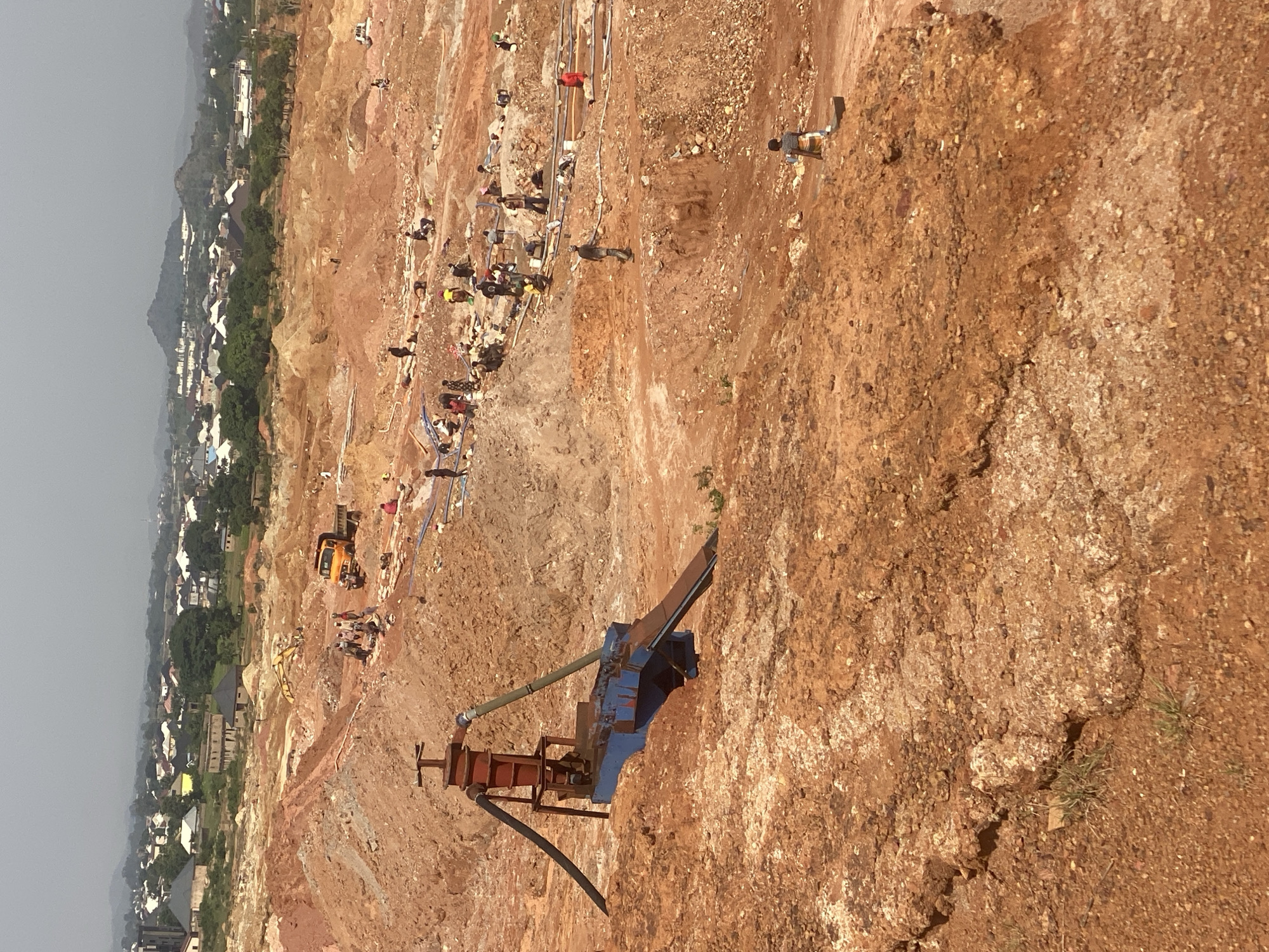 The image depicts an open-pit mine in Jos, Nigeria, characterized by vast expanses of disturbed earth with reddish-brown soil and scattered rocks. The terrain is uneven, with steep slopes and mounds of excavated material. In the foreground, a piece of mining equipment, possibly a sluice box or a concentrator, sits partially submerged in the soil. It has a blue metal structure with a reddish-orange hopper or feeder mechanism. Hoses, one black and one green, are connected to the equipment.
In the midground and background, numerous people are engaged in mining activities. They are scattered across the landscape, some working in groups, others individually. Their clothing is varied, with some individuals wearing brightly colored attire. Several people appear to be digging, carrying materials, or operating smaller pieces of equipment. A yellow truck is visible in the middle distance, suggesting the transport of excavated materials. In the far background, the city of Jos is visible, with residential buildings and a range of hills under a hazy sky. The overall scene suggests an active and labor-intensive mining operation, with the urban environment of Jos forming a backdrop.