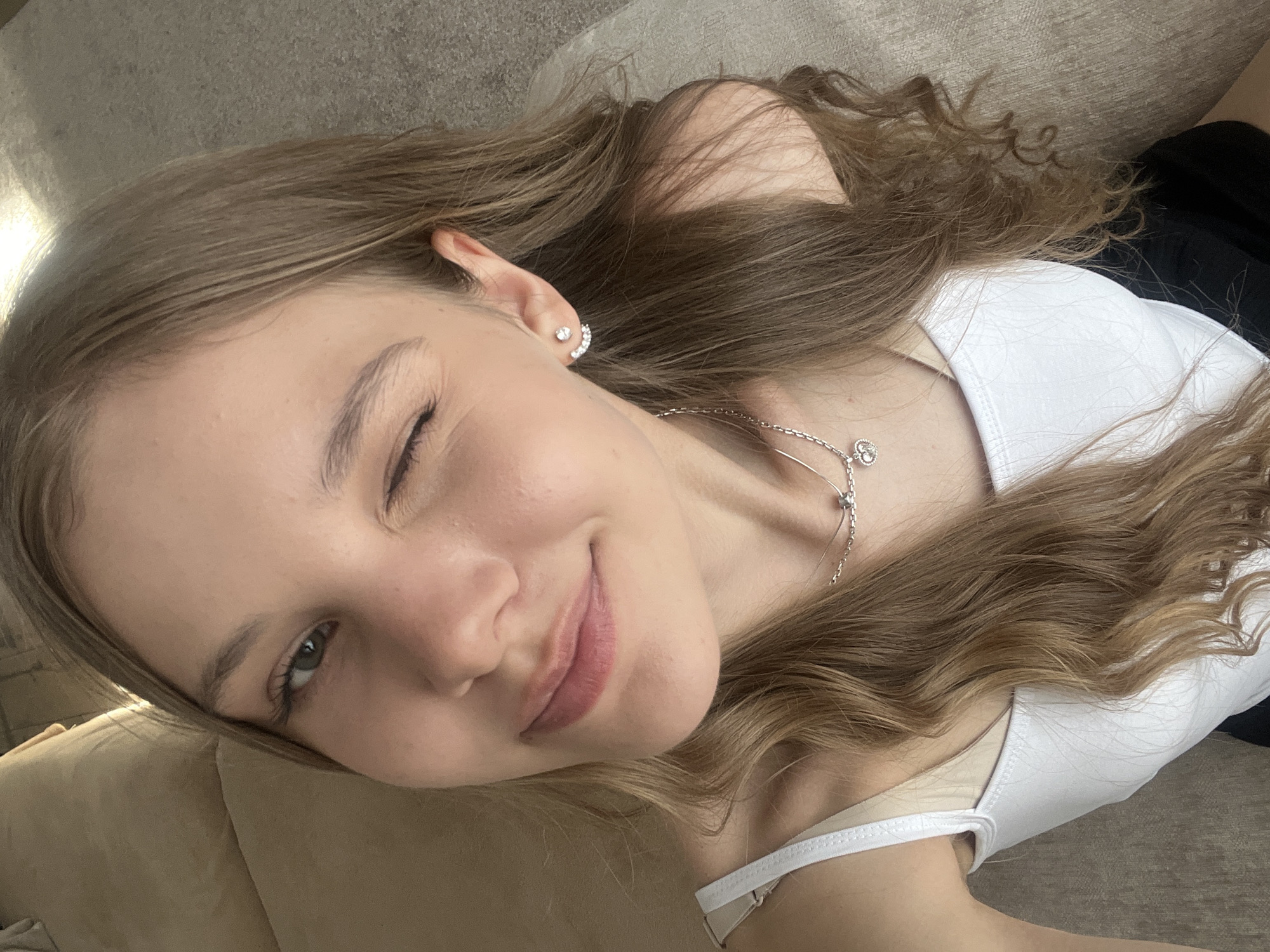 A young woman lies on a beige couch, winking at the camera. She has long, wavy brown hair and is wearing a white tank top and a silver necklace. Her left ear is adorned with a sparkly stud earring. The location is not discernible as being in Votkinsk, Russia or any specific city.