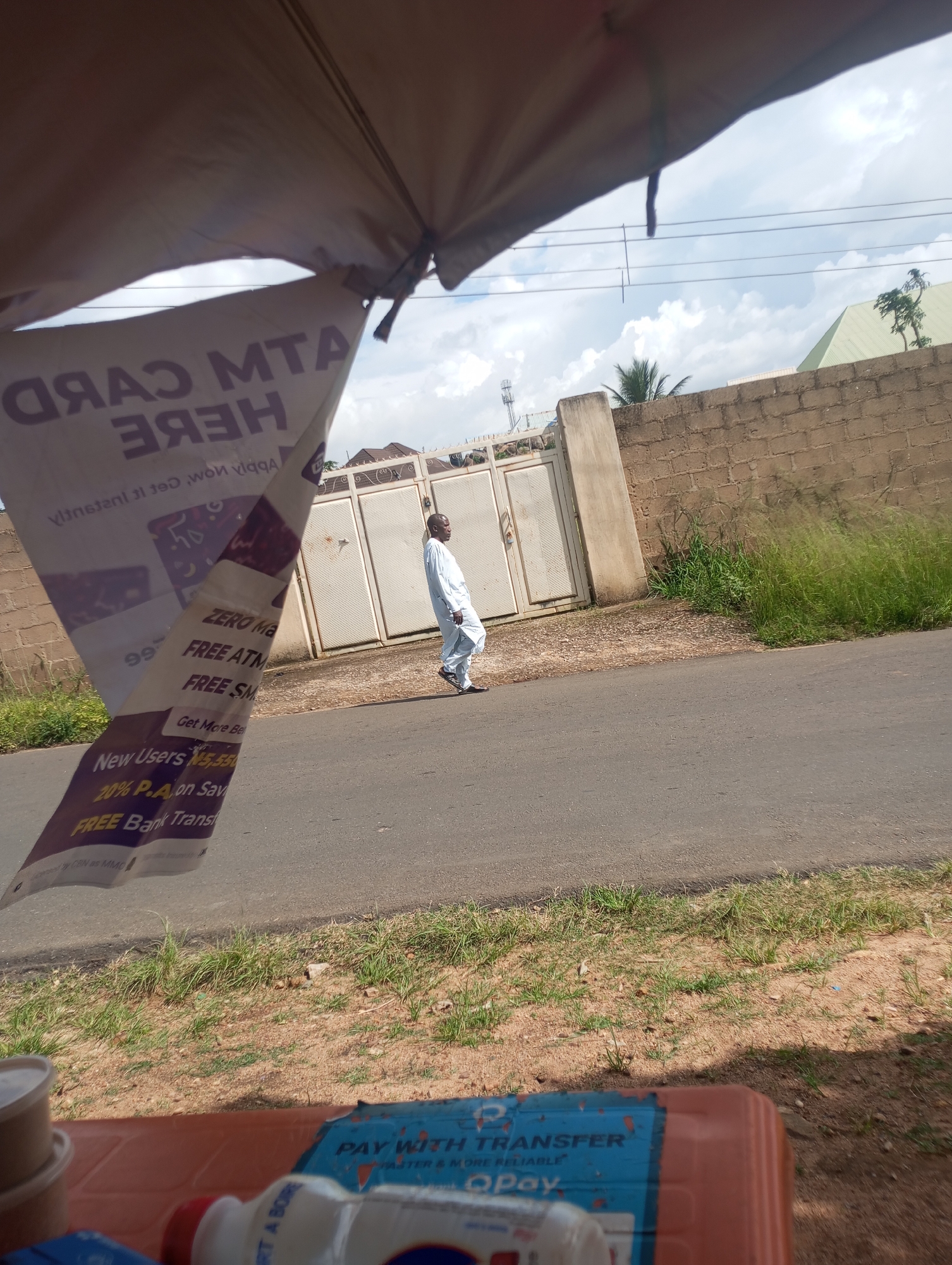 An outdoor medium shot captures a man walking along a paved road. He is wearing a white traditional outfit and black sandals. The man is walking away from the camera towards a gated compound with a brick wall surrounding it. To the left of the frame, a sign with text is hanging, partially obscured by the canvas of an outdoor shelter. The sign advertises "ATM CARD HERE," "ZERO MA," "FREE ATM," "FREE SM," "New Users 20% P.A on Sav," and "FREE Bank Trans." In the foreground, a blurred image shows part of a blue and orange container, suggesting a market or roadside stall. The sky is partly cloudy.