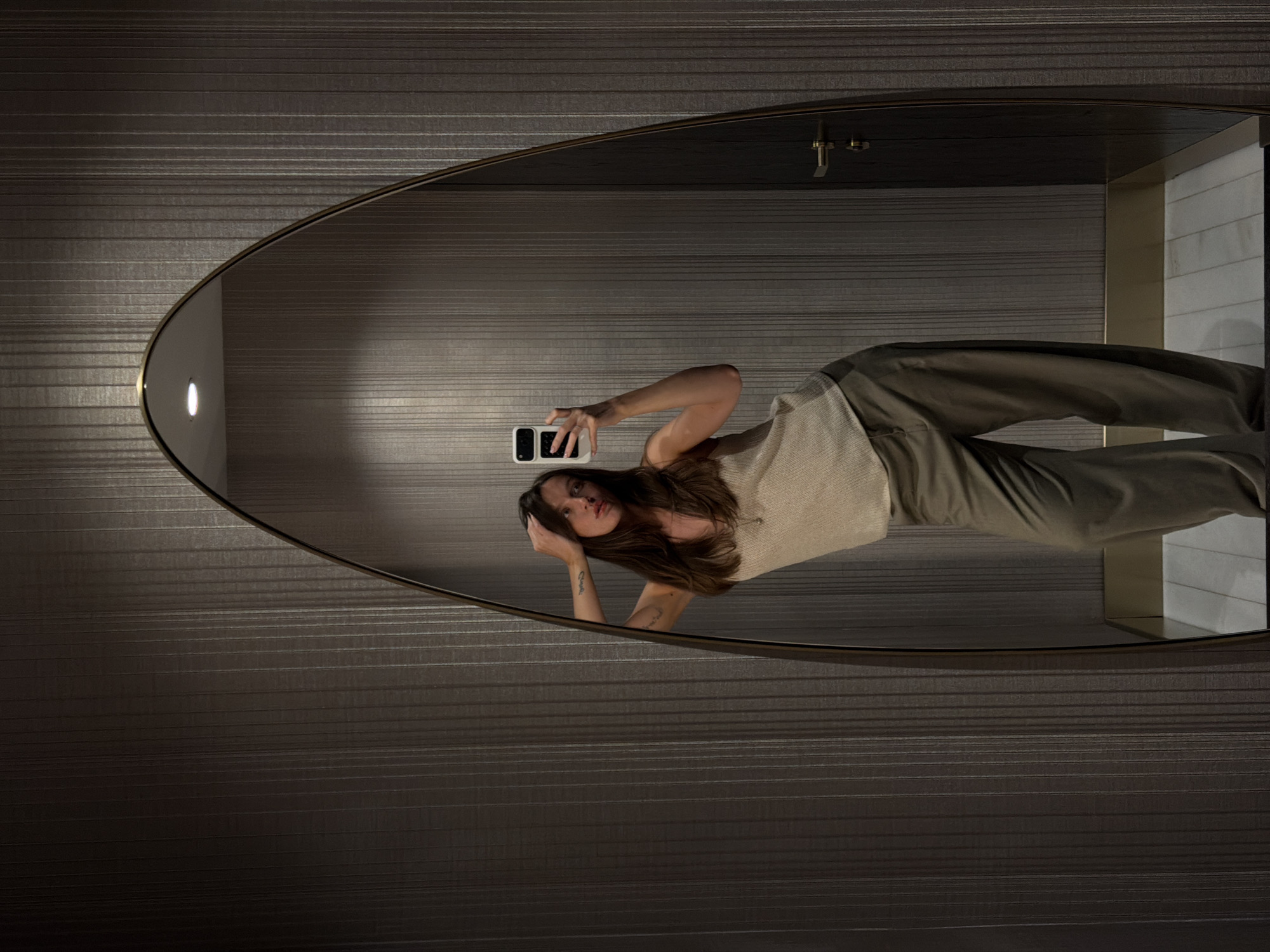 A woman stands in front of an oval mirror, holding a phone up to take a selfie. The mirror reflects the woman, who is wearing a light-colored top and olive-green pants. The background consists of a wall with horizontal striped texture. The scene appears to be indoors, likely in a bathroom or dressing area. The image doesn't provide sufficient information to confirm it was taken in Budva, Montenegro.