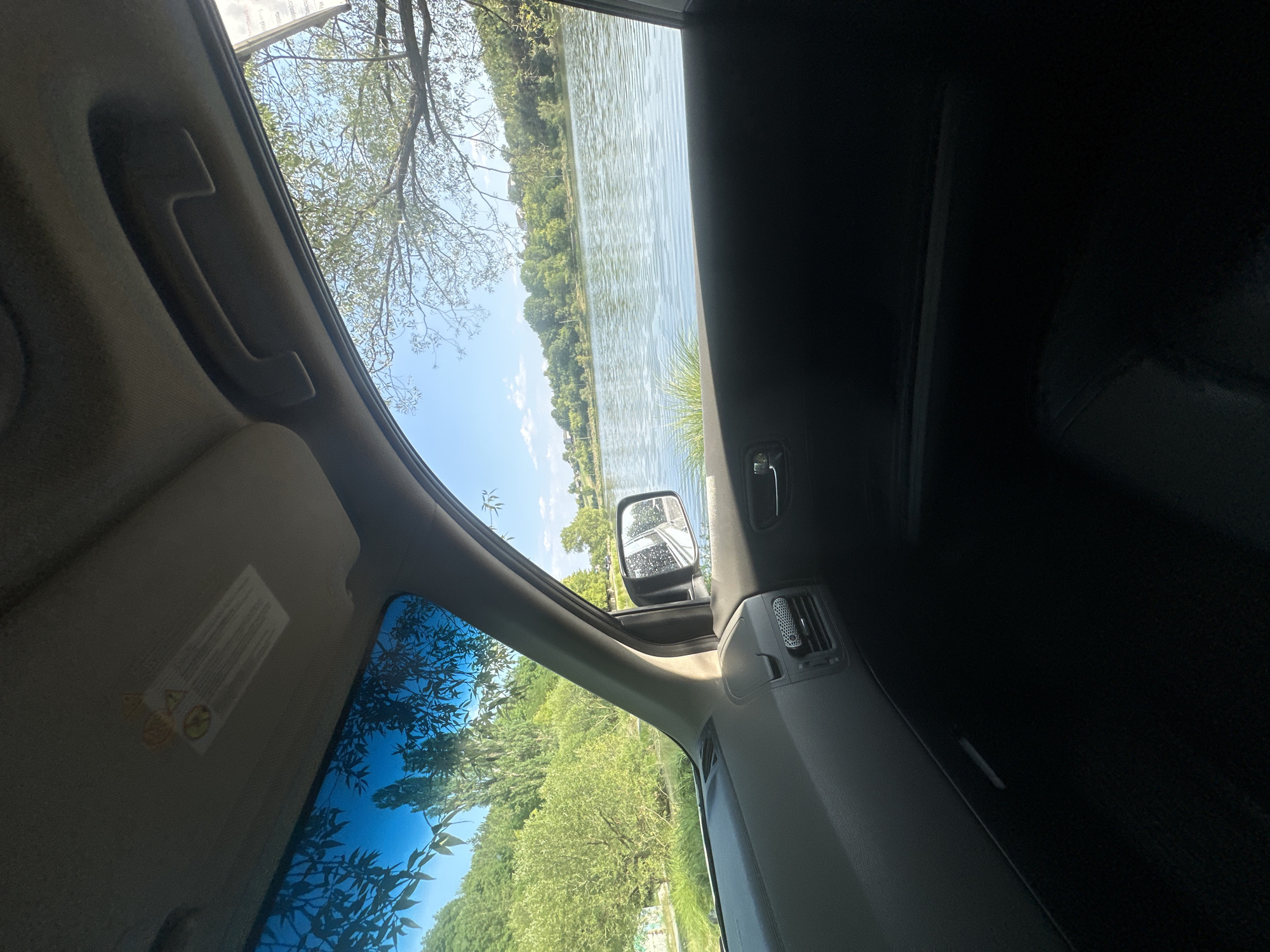 The image shows a view from inside a car looking out towards a body of water and a tree-lined landscape. The car's interior is visible in the foreground, with the dashboard, a side mirror, and part of the door. The side mirror reflects a road and trees. Outside the car, a lake or river is prominent, with green vegetation and trees along its banks. The sky is blue with some scattered clouds. The overall scene appears to be a tranquil outdoor setting. There are no people visible in the image. The image is a photograph taken during the day, likely on a sunny day, given the bright lighting and clear sky. The car appears to be parked near the water's edge.
The location of Makiv, Ukraine is not identifiable from the image itself, as there are no specific landmarks or identifying features.