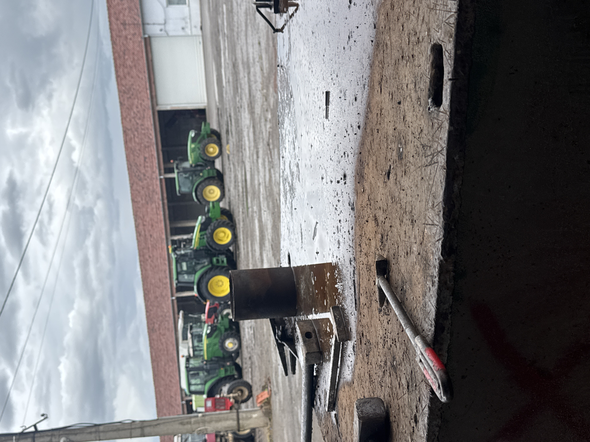 The image shows a partially obscured foreground with various tools and metal objects, including a pipe and a hammer, resting on a wet, possibly concrete, surface. In the background, a line of green and yellow John Deere tractors are parked in front of a large brick building with metal doors. The sky is overcast. There is no direct evidence to confirm that the location is in the city of Sarnevo, Bulgaria. There are no people visible in the image. The interaction between the objects seems to be one of storage or temporary placement. The environment suggests an outdoor workspace, possibly a farm or agricultural service area.
