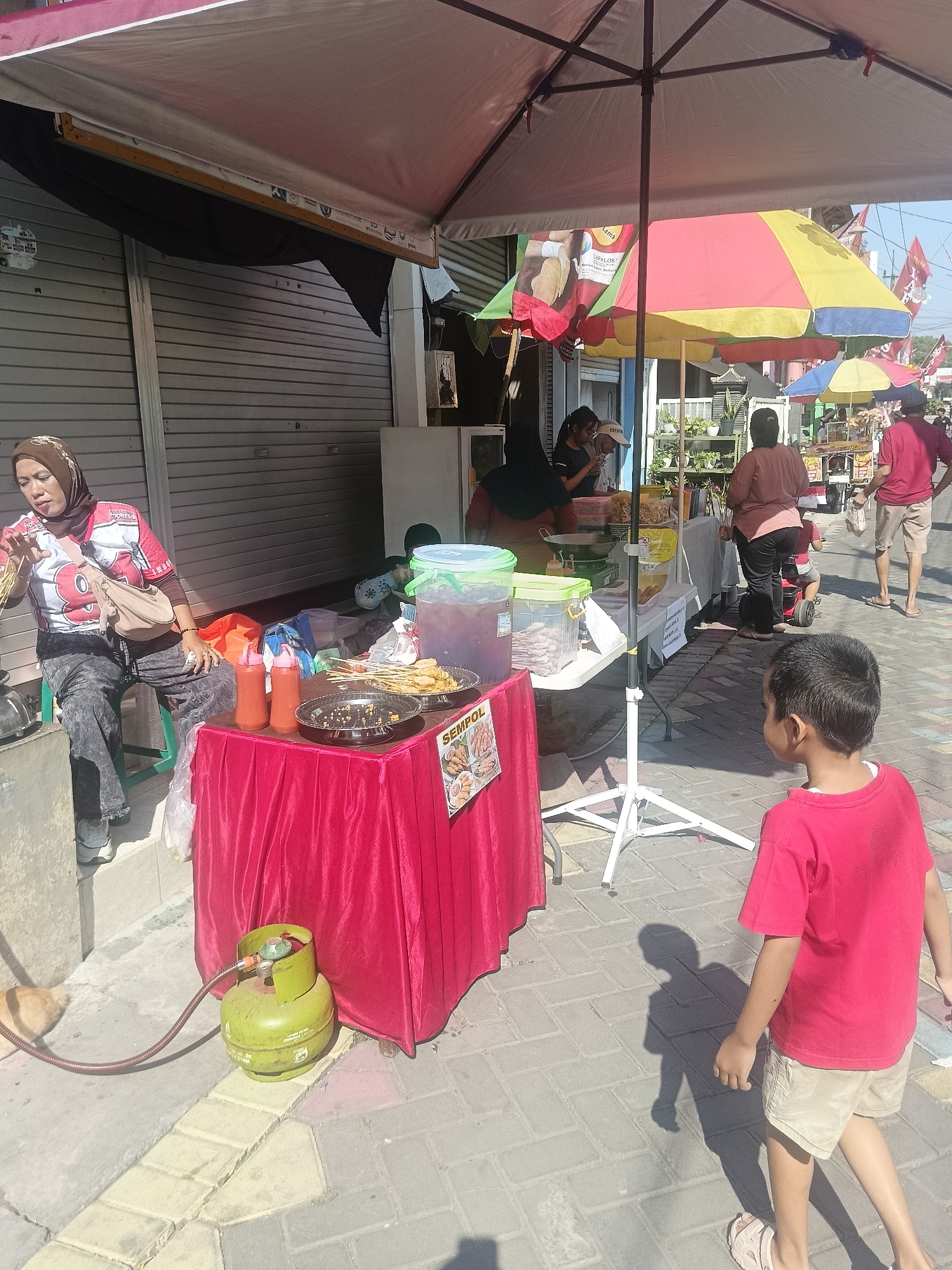 This image depicts an outdoor street scene in Gedangan, Indonesia, likely a market or food stall area. A woman wearing a hijab and a red and white shirt is seated on a green stool, preparing food at a stall covered by a large pink and yellow umbrella. In front of her, a table draped with a red cloth displays food items, including skewers and bottles of sauce. A green gas cylinder is positioned on the ground next to the stall, connected by a hose.
Several other individuals are present in the background. To the right of the woman, a young boy in a red shirt and beige shorts is walking away from the camera. Further back, other people are seen interacting at different stalls, some under colorful umbrellas. One stall appears to be selling drinks in large containers. The ground is paved with interlocking bricks. The overall atmosphere suggests a bustling commercial environment during daylight hours.