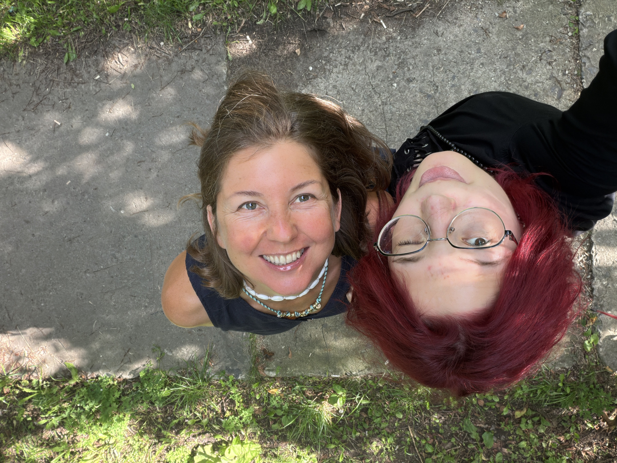 The image shows a high-angle, close-up selfie of two women outdoors. The woman on the left has bright red hair and is wearing glasses and a black hooded jacket. She is winking and sticking her tongue out. The woman on the right has brown hair and is wearing a black t-shirt and a beaded necklace. She is smiling broadly, showing her teeth. They appear to be standing on a paved surface next to an area of grass. The lighting suggests it is daytime, with dappled sunlight. The background consists of grass and patchy soil. The specific location in Chelyabinsk, Russia, cannot be determined from the image alone.