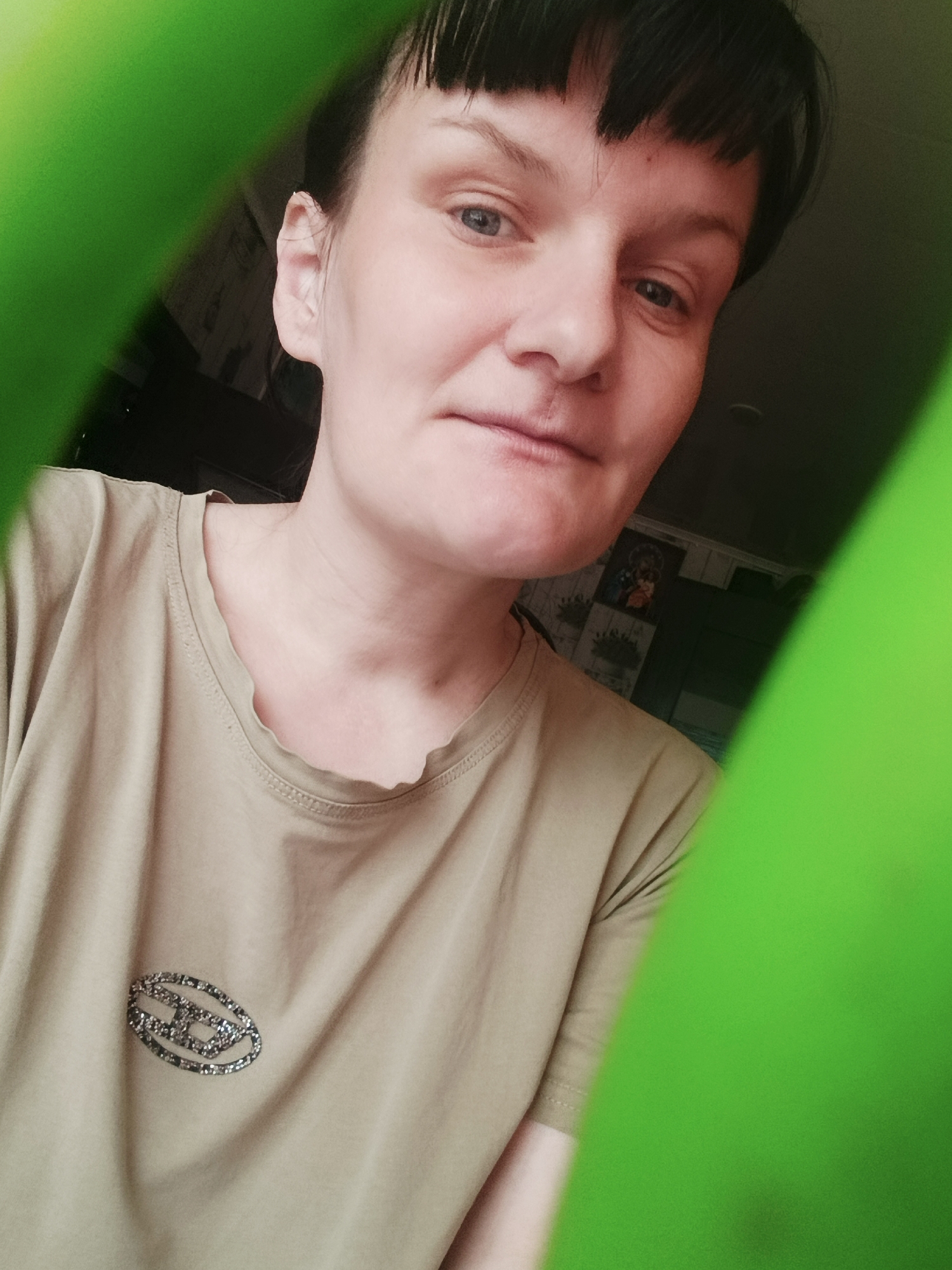 A close-up portrait shows an adult individual with fair skin, dark short hair, and bangs, looking directly at the camera with light-colored eyes and a neutral expression. The person wears a light olive green or khaki-colored short-sleeved t-shirt with a slightly ruffled neckline. On the left side of their chest, a distinctive oval-shaped emblem is visible, made of sparkling, possibly silver or grey, embellishments that form a stylized design or intertwining letters.
The image is framed by two out-of-focus, bright green objects on the left and right edges, partially obscuring the view. These objects create a soft, tunnel-like effect around the central subject. The background behind the person is dark and blurred, suggesting an indoor setting. Faint details like what appears to be framed pictures or decorations on a wall, and a dark furniture piece or wall section, are subtly visible behind their head and right shoulder. The overall impression is a casual, intimate selfie or portrait taken in a domestic environment. There is no clearly decipherable text visible, only the abstract emblem on the shirt.