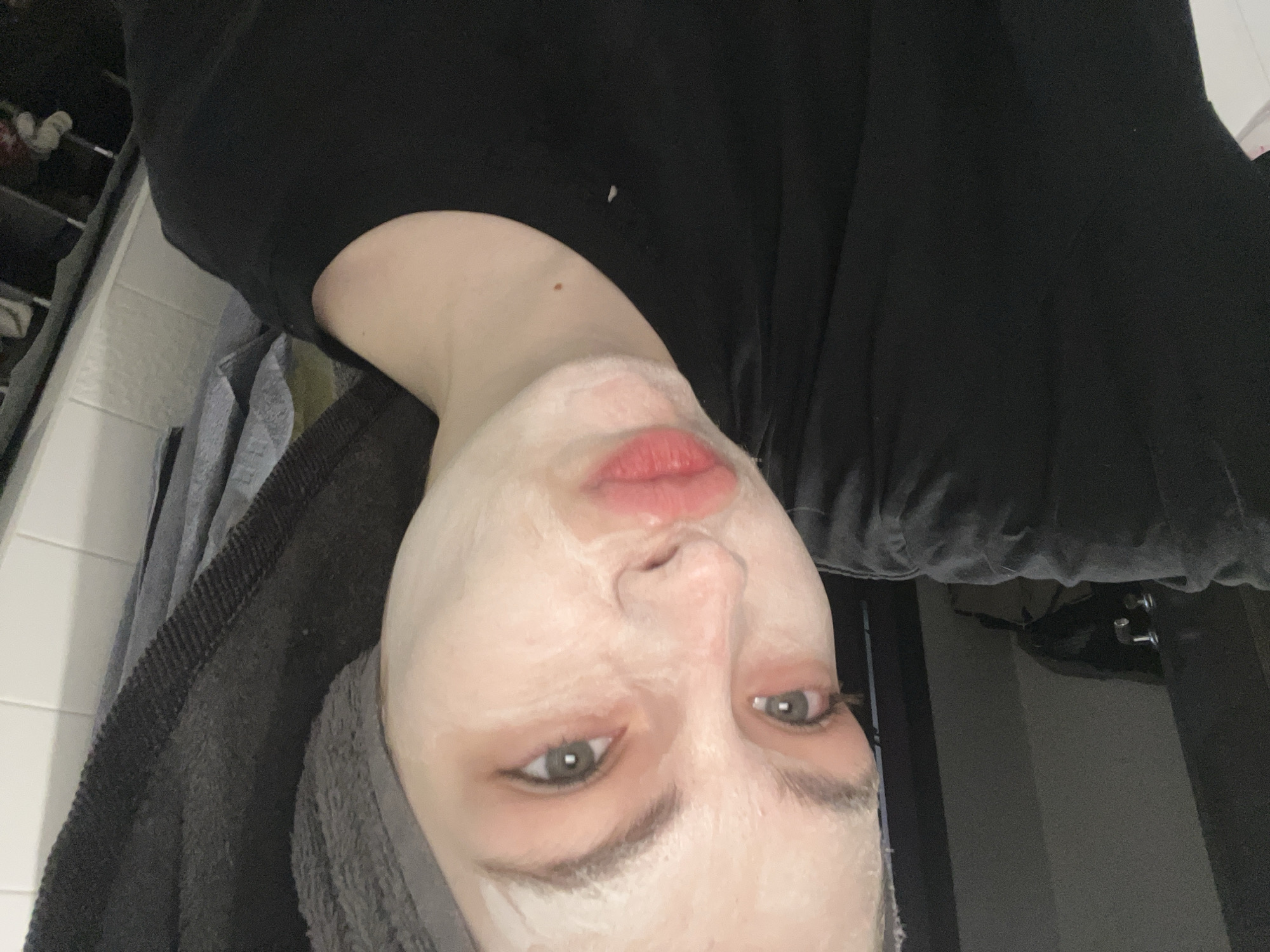 A person with a face mask is positioned at an inverted angle, facing directly toward the camera. The person has gray-green eyes and fair skin. Their lips appear red, possibly due to lip color or product application. A gray towel is draped along the left side of the frame. The person is wearing a black shirt. A shelf unit is partially visible in the background on the left, while a section of a gray wall and a towel bar are visible on the right. The image was not taken in Saint Petersburg, Russia.