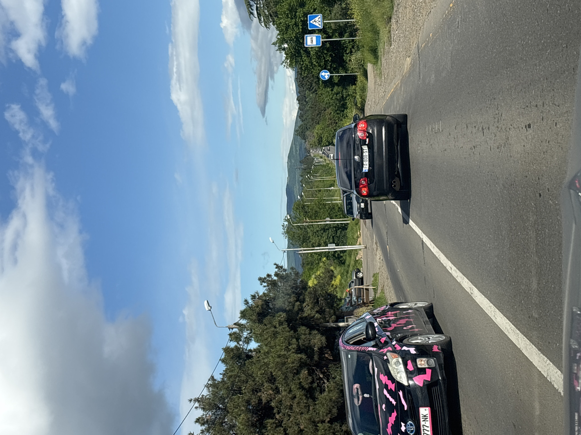 The image depicts a road scene with a view of mountains in the distance, suggesting a location on the outskirts of a city. The sky is blue with scattered clouds. In the foreground, there are several cars. One car, in the foreground, is black with pink decals and a license plate. Behind it is a black car with license plate FA-808-ST. The road is paved with a white line marking the lanes. Road signs are visible on the side of the road. The scene appears to be Tbilisi, Georgia, based on the license plates, road signs, and landscape.