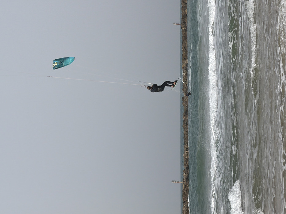 A coastal scene featuring kiteboarding near the Jaffa Port breakwater in Tel-Aviv, Israel. An individual, wearing a dark wetsuit, is captured airborne in a dynamic kiteboarding maneuver, suspended above the water. The kiteboarder holds a control bar connected via lines to a teal-colored North brand kite visible in the upper right. Their feet are strapped to a dark kiteboard, also elevated above the water. A second, partially submerged figure or object is indistinctly visible in the water directly beneath the airborne board. The foreground shows shallow ocean water with small breaking waves approaching a light-colored sandy beach. In the mid-ground, a substantial rock breakwater spans the width of the frame, featuring two navigation markers: a red-and-white striped pole with a red top-mark on the left, and a less distinct pole on the right. The sky is overcast, presenting a uniform light grey-blue hue over the entire scene.