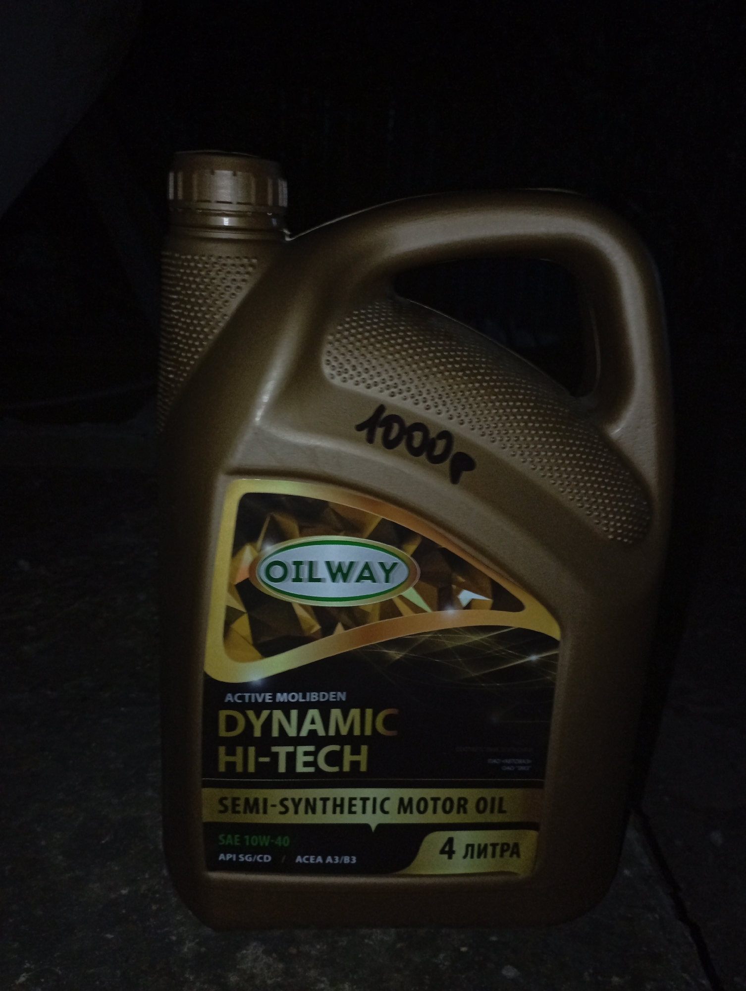 The scene, located in Kurganinsk, Russia, depicts a single gold-colored 4-liter plastic container of "OILWAY DYNAMIC HI-TECH SEMI-SYNTHETIC MOTOR OIL" standing upright in a dimly lit environment. The container features an integrated handle on the right side and a ribbed screw cap on the top left. A black marker inscription, "1000P," is visible on the upper front surface of the container, to the left of the handle. The main label displays "ACTIVE MOLIBDEN," "SAE 10W-40," "API SG/CD," and "ACEA A3/B3" specifications against a gold and black geometric background. The container rests on a dark, textured surface, possibly concrete or pavement, which exhibits a diagonal fissure or seam in the lower right portion of the frame. The background is uniformly dark and indistinct. No individuals or active subjects are present.