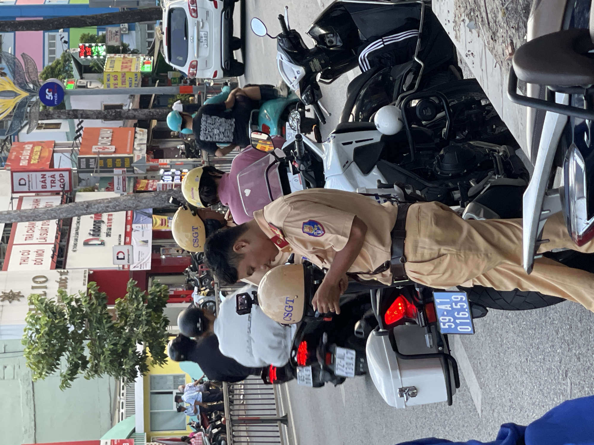 The image shows a Vietnamese traffic police officer in Thủ Đức, Vietnam inspecting a parked motorcycle. The officer is wearing a tan uniform and appears to be examining the motorcycle's license plate. Several other motorcycles are parked in a row nearby, along with pedestrians. Buildings with signs and advertisements line the street. The license plate on the motorcycle is visible, displaying the number "59-A1 016.59". Other signs with OCR text include 72-AL 139.47, HELIO ĐỔI MỚI LÃI SUẤT and ONE WAY. The text CSGT is visible on the helmets.
