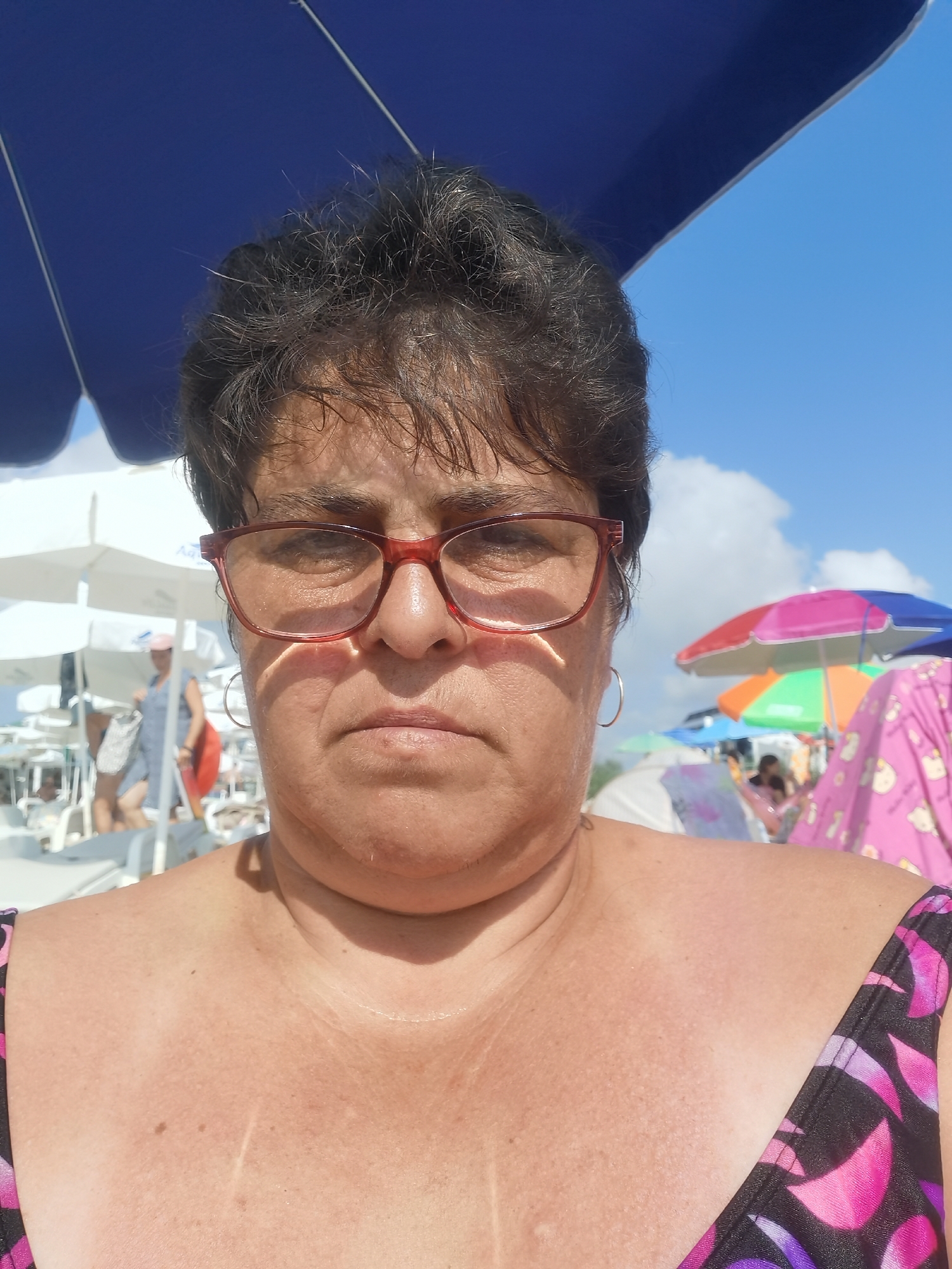 A close-up, front-facing selfie shows a woman with dark, short hair and glasses on a beach. She is wearing a dark swimsuit with a pink and purple floral pattern. The background suggests a sunny day with a blue sky, beach umbrellas, and people in the distance. The overall setting is a beach resort.