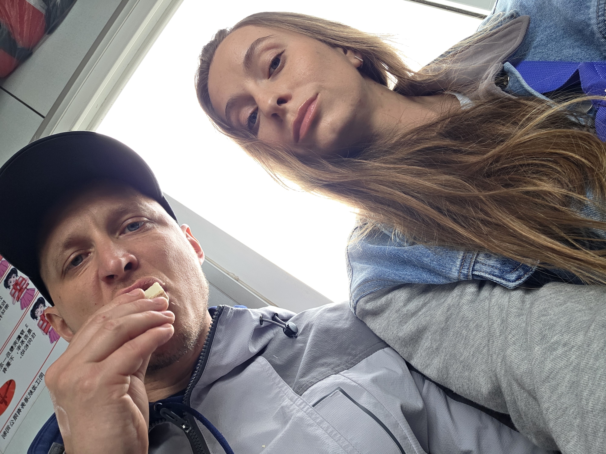 This low-angle selfie captures two individuals, a man and a woman, looking towards the camera against a bright white background, likely an overhead light source or window.
On the left, a man is seen from the chest up, wearing a black baseball cap and a grey and blue zip-up jacket. He has light stubble and blue eyes. He holds a small, light-colored piece of food (resembling cheese or a biscuit) between his thumb and index finger, bringing it towards his mouth as if about to eat. Adjacent to him, on the far left, a white panel displays red and black text, clearly identifiable as Chinese characters, alongside stylized, cartoon-like illustrations of people, possibly women in uniform or traditional attire.
On the right, a woman is visible from the chest up, her long, light brown hair flowing over her shoulder. She wears a light blue denim jacket over a grey long-sleeved top. She looks directly at the camera with a calm and neutral expression, her face slightly angled.
The bright background and the indoor-like setting suggest the image was taken inside a vehicle or building. The Chinese text on the wall serves as a significant location cue. The overall atmosphere is casual, capturing a shared, candid moment.