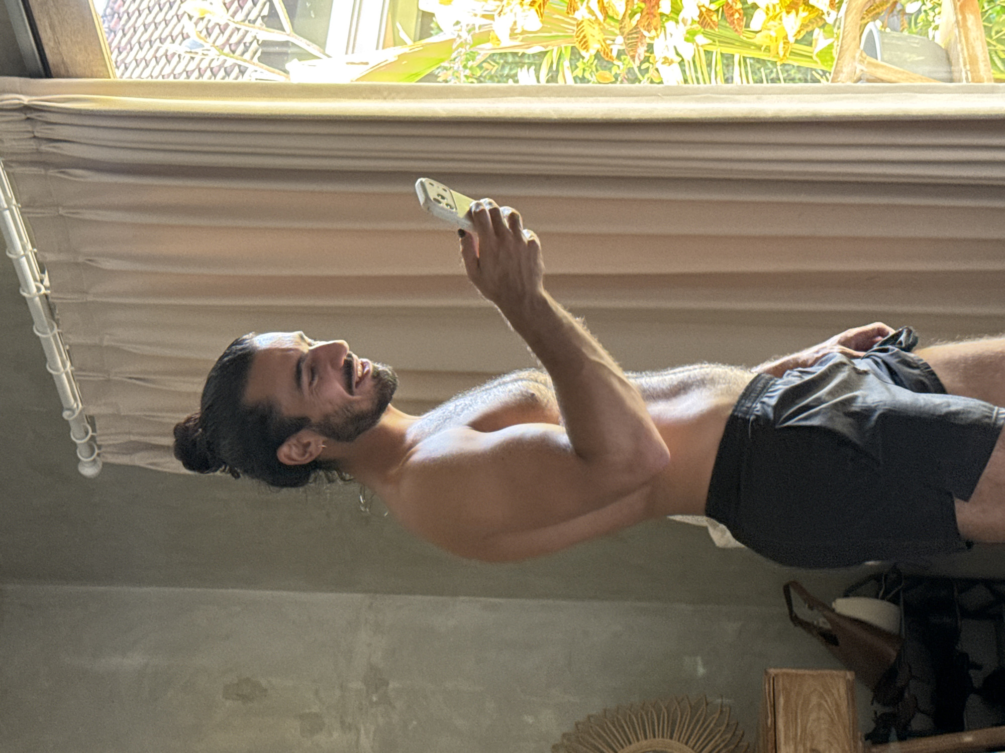 In North Kuta, Indonesia, a shirtless man with his hair in a bun stands facing right, looking at his phone. He is wearing black shorts. Sunlight streams in from a window to his right, illuminating the room and highlighting a beige curtain and lush greenery outside. To his left, a textured grey wall and wooden furniture are visible.